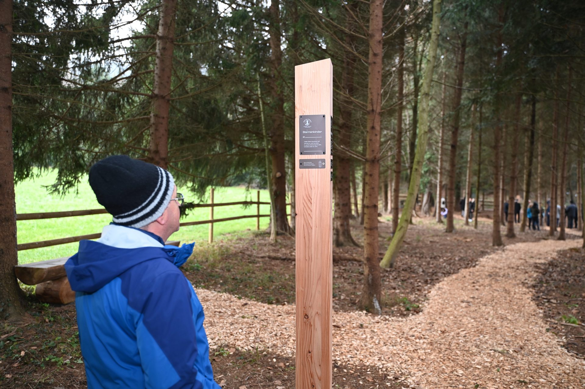 Man in blue jacket and beanie looks at a wooden signpost in a forest with a path and people.