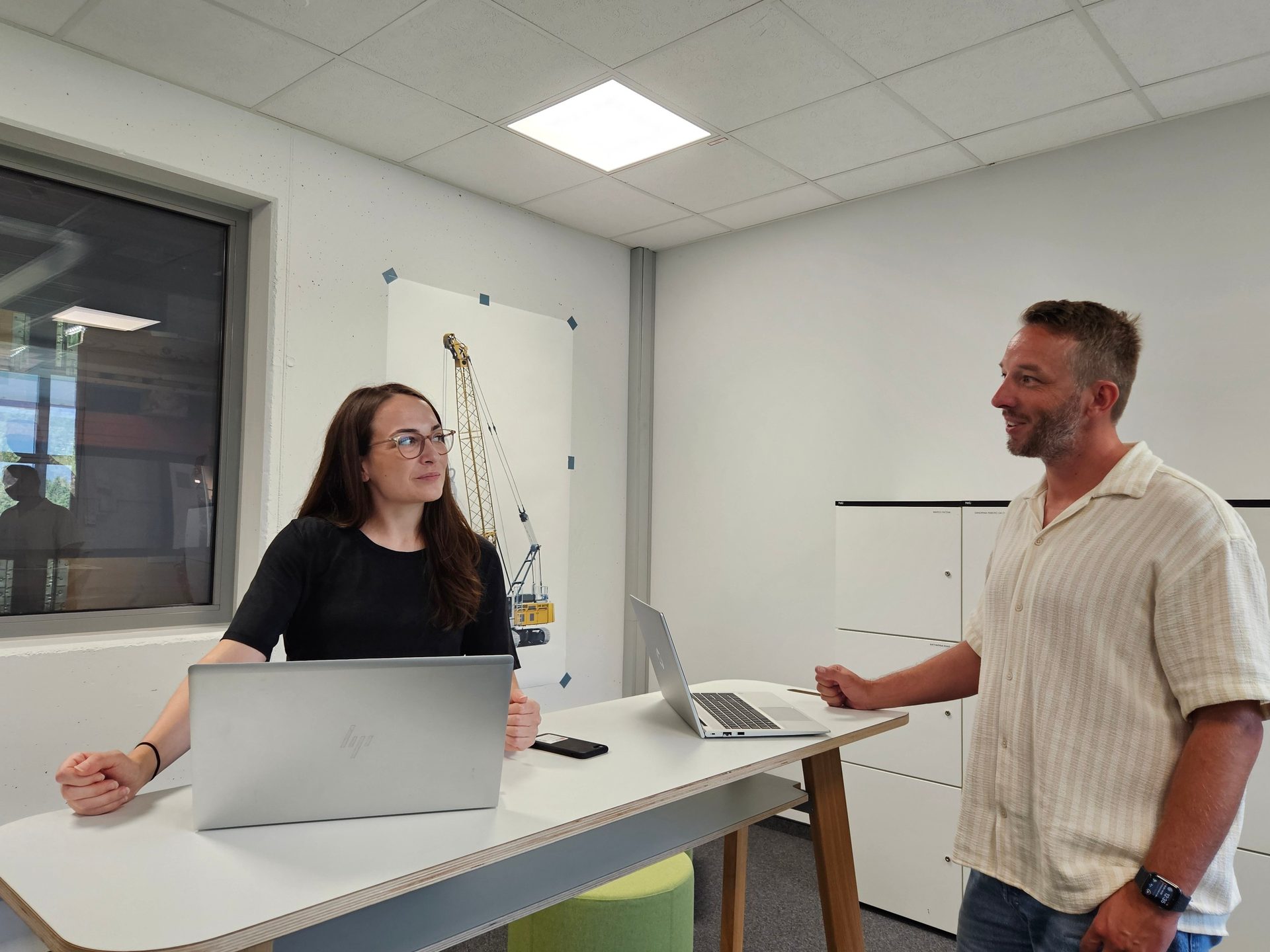 Two colleagues in an office with laptops, having a discussion.