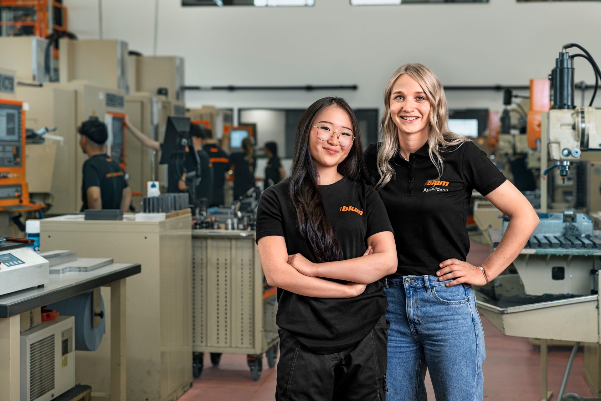 Two smiling women, one East Asian, one white, in black Blum shirts, pose in a busy factory.