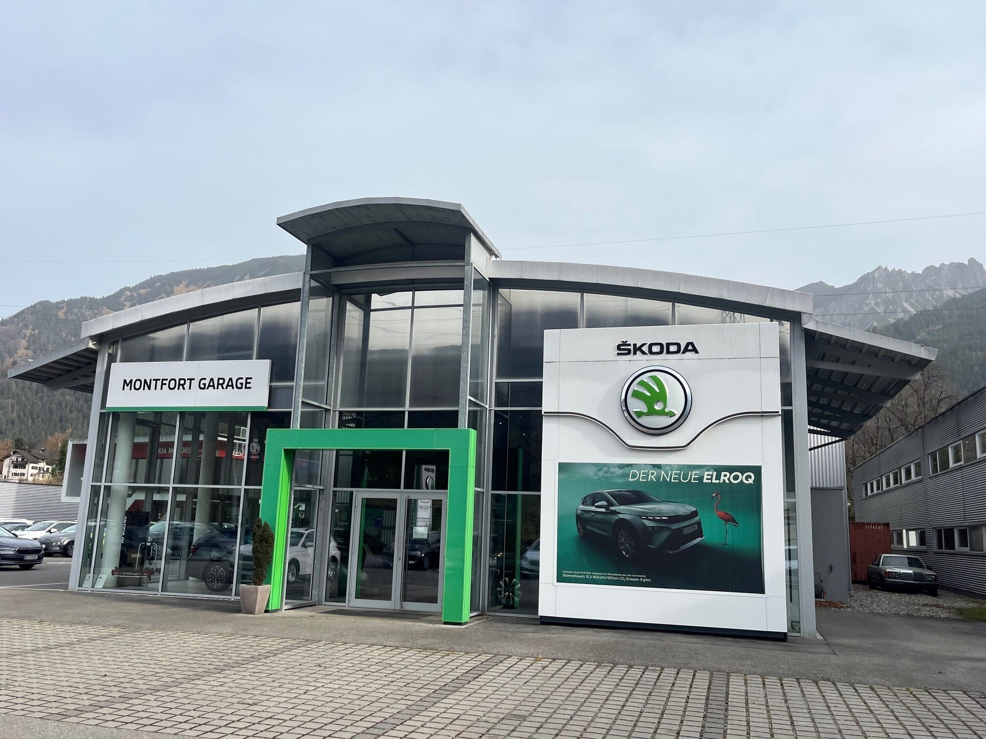 Montfort Garage car dealership with Skoda branding and "DER NEUE ELROQ" ad. Mountains in background.