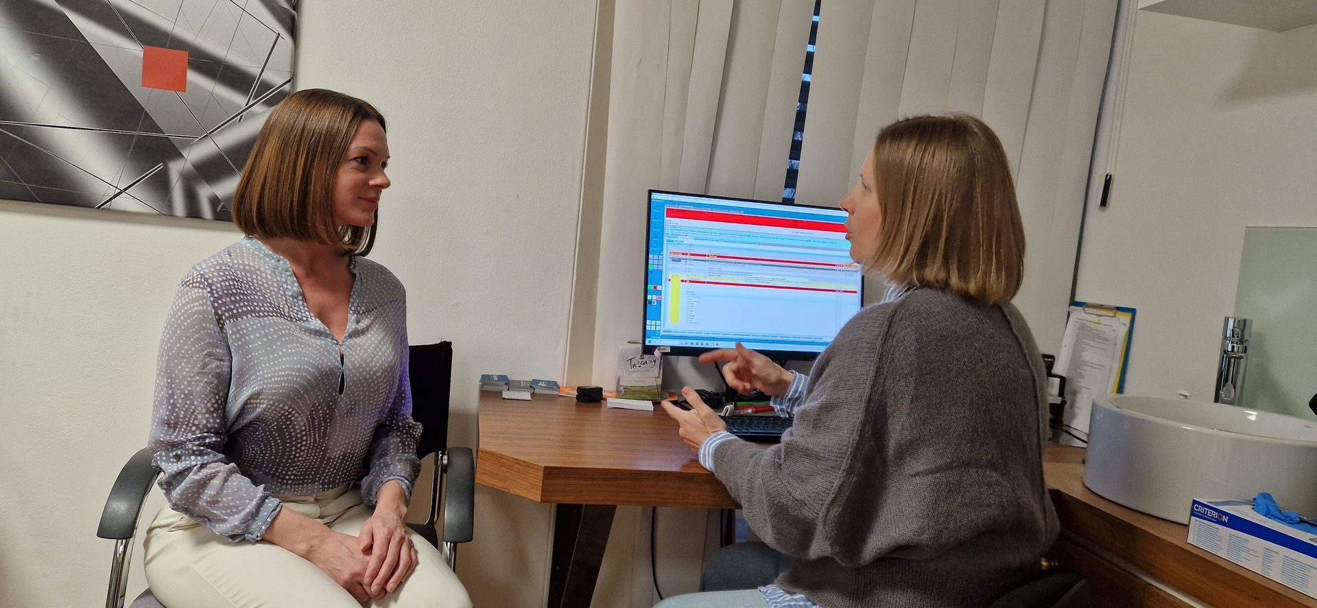 Two women in a room, one seated, the other at a desk with a computer screen.