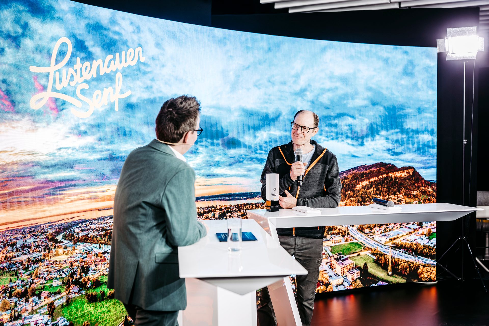 Two men on a stage, one holding a microphone, in front of a large screen displaying "Lustenauer Senf" and a landscape.