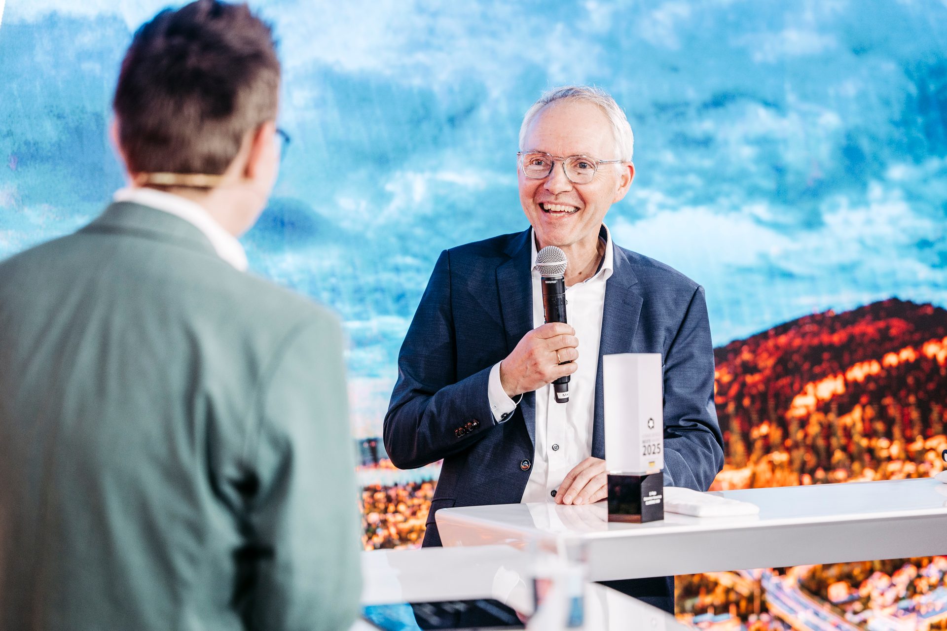 Smiling man in suit holds a microphone, speaking to another person at an event with a scenic backdrop.