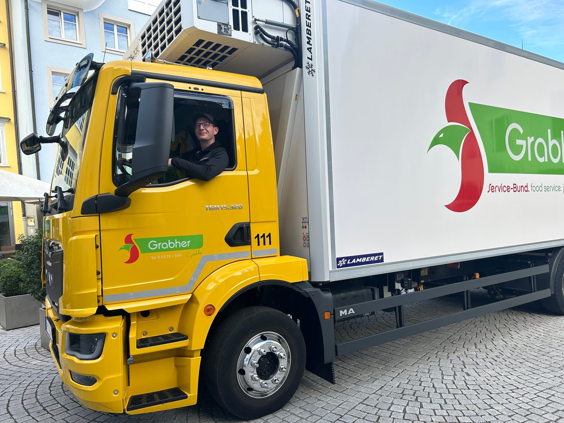 A man smiling from the driver's seat of a yellow Grabher delivery truck.