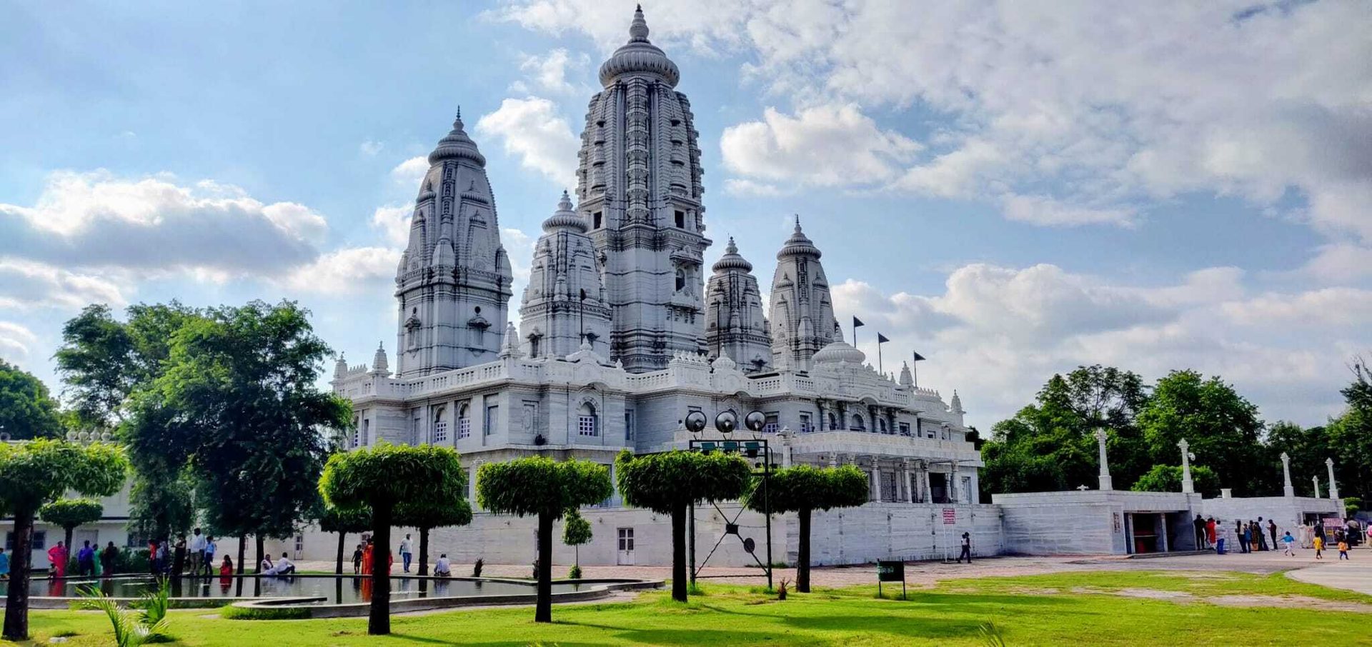 A grand white temple with multiple towers, surrounded by green trees and grass, under a cloudy blue sky.