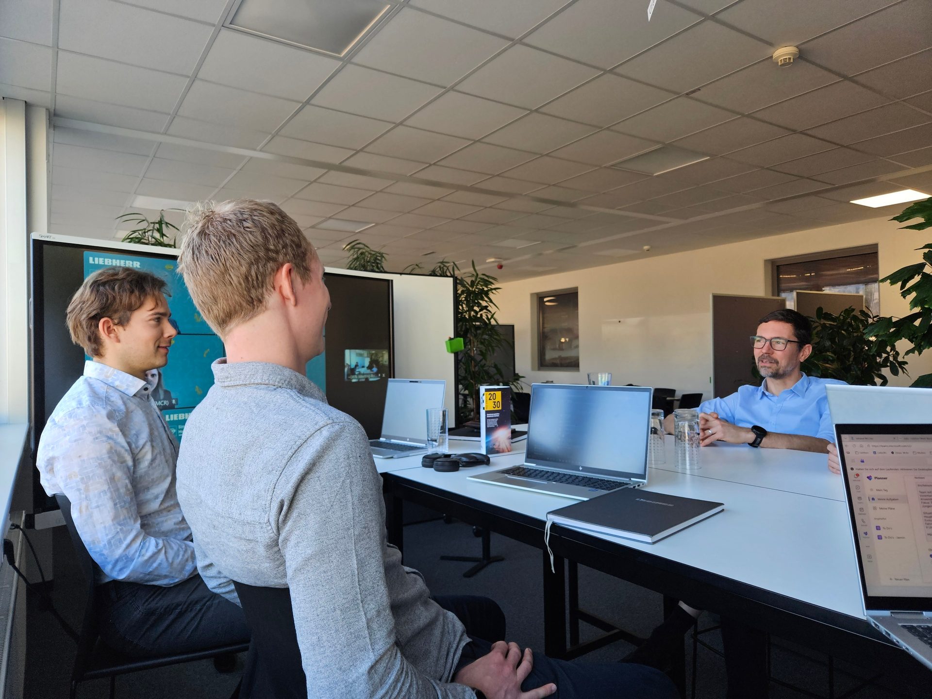 Three men in a modern office, two observing a screen, one speaking during a business meeting.