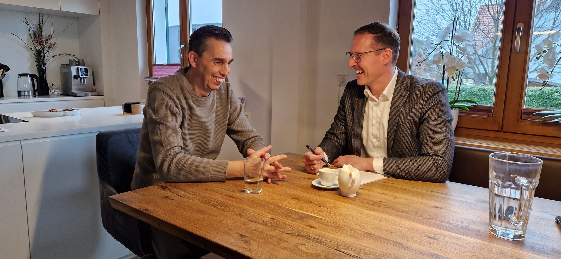 Two laughing men having a conversation at a wooden table with coffee, water glasses, and kitchen in background.