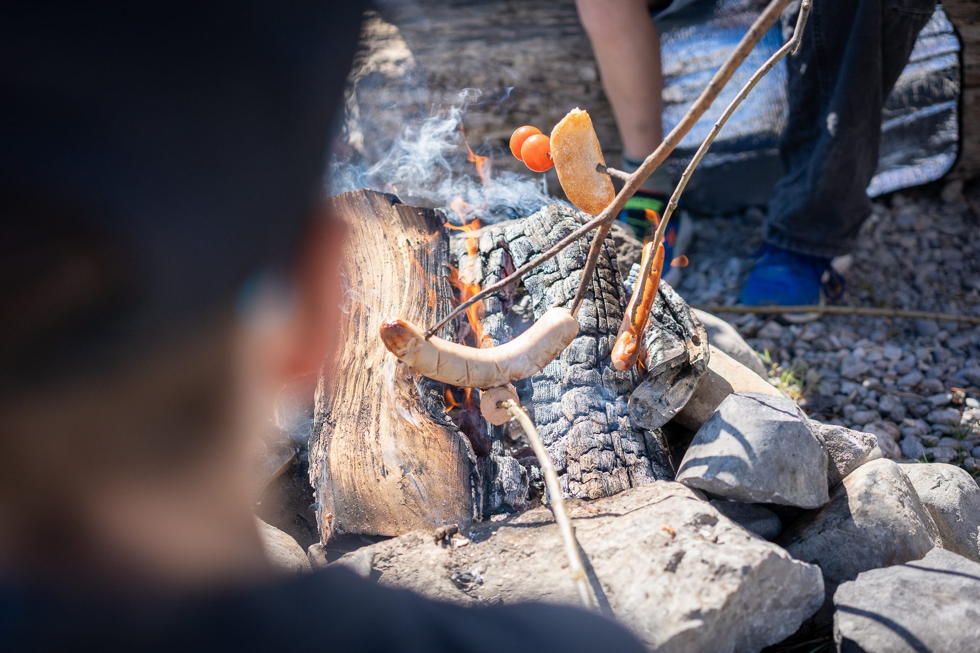 Close-up of people roasting sausages, bread, and tomatoes over a rustic campfire.