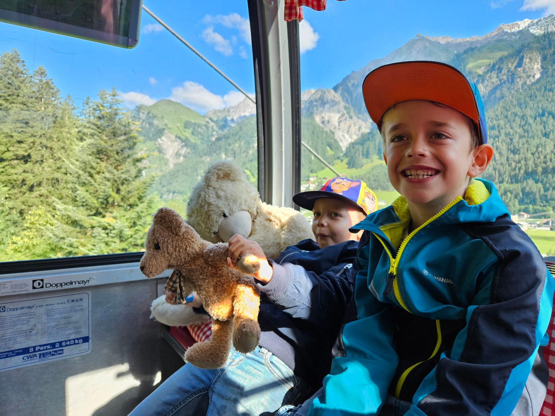 Two smiling boys and teddy bears inside a cable car overlooking mountains.