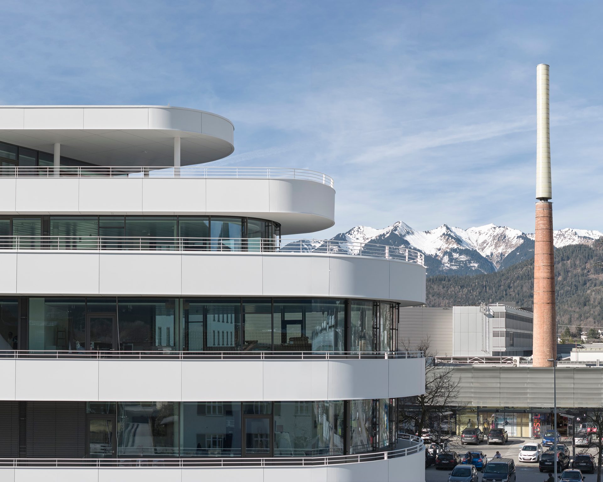 Modern white building with curved balconies, a tall brick chimney, and snow-capped mountains under a blue sky.