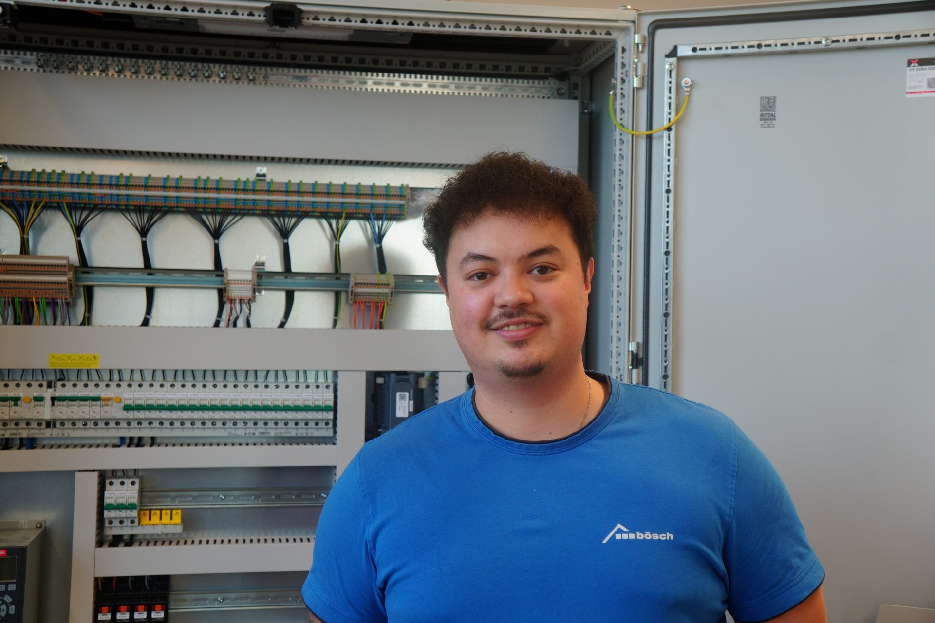 Young man in a blue shirt smiles in front of an open electrical control cabinet with complex wiring.