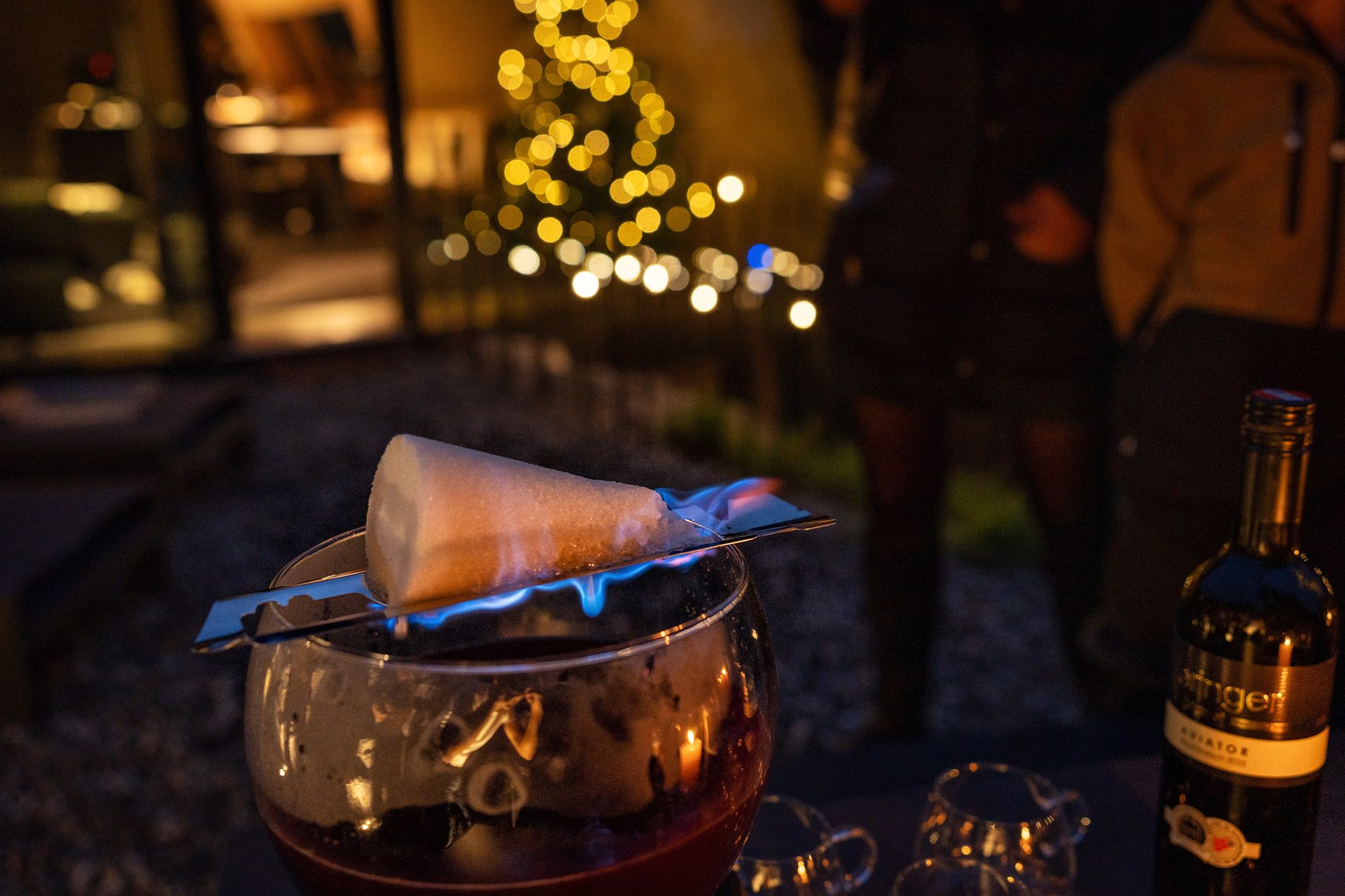 A flaming sugar cone melts into a glass of punch, with festive lights and people in the background.