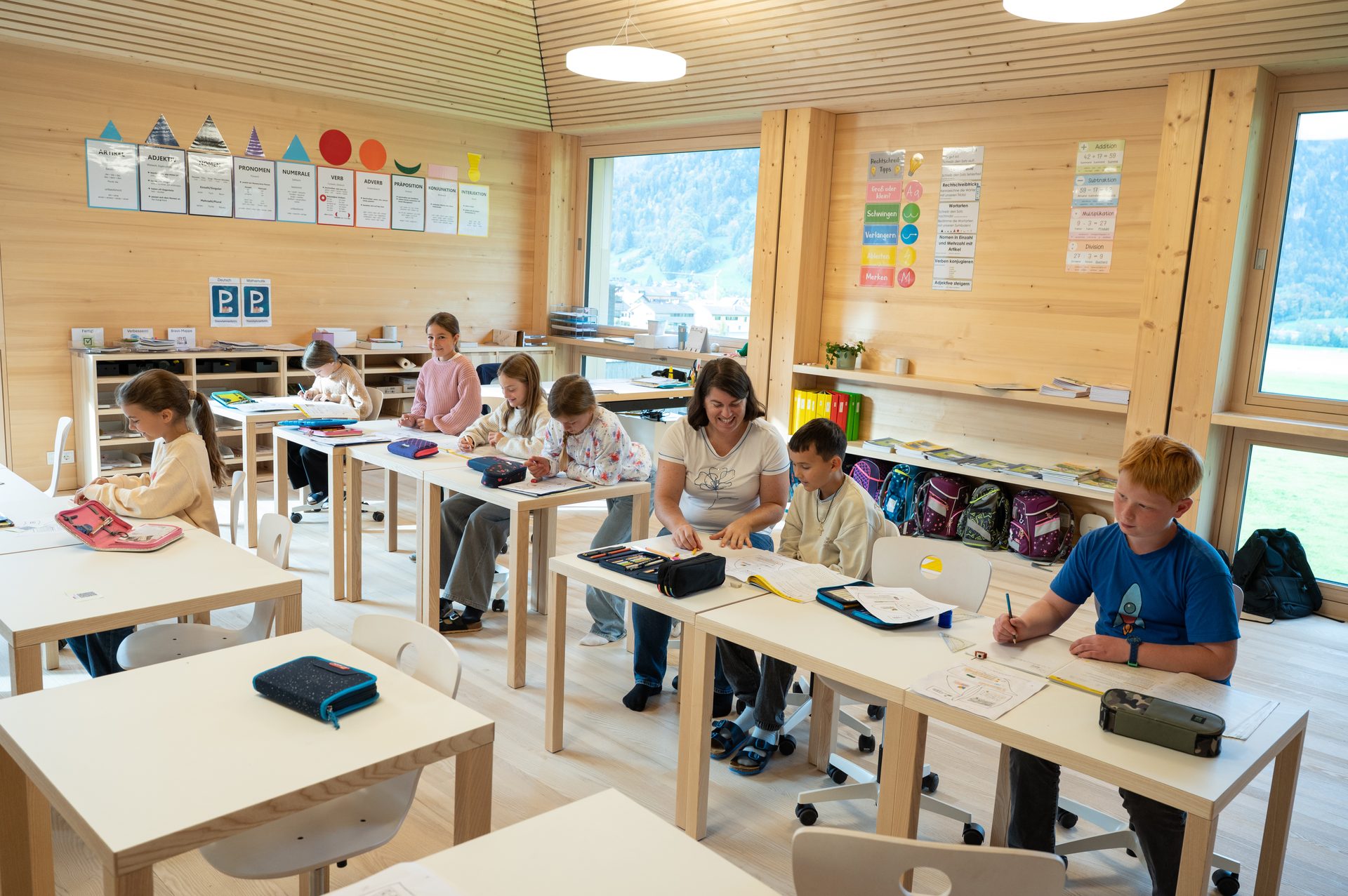 A bright classroom with children and an adult working at wooden desks, educational posters on walls.