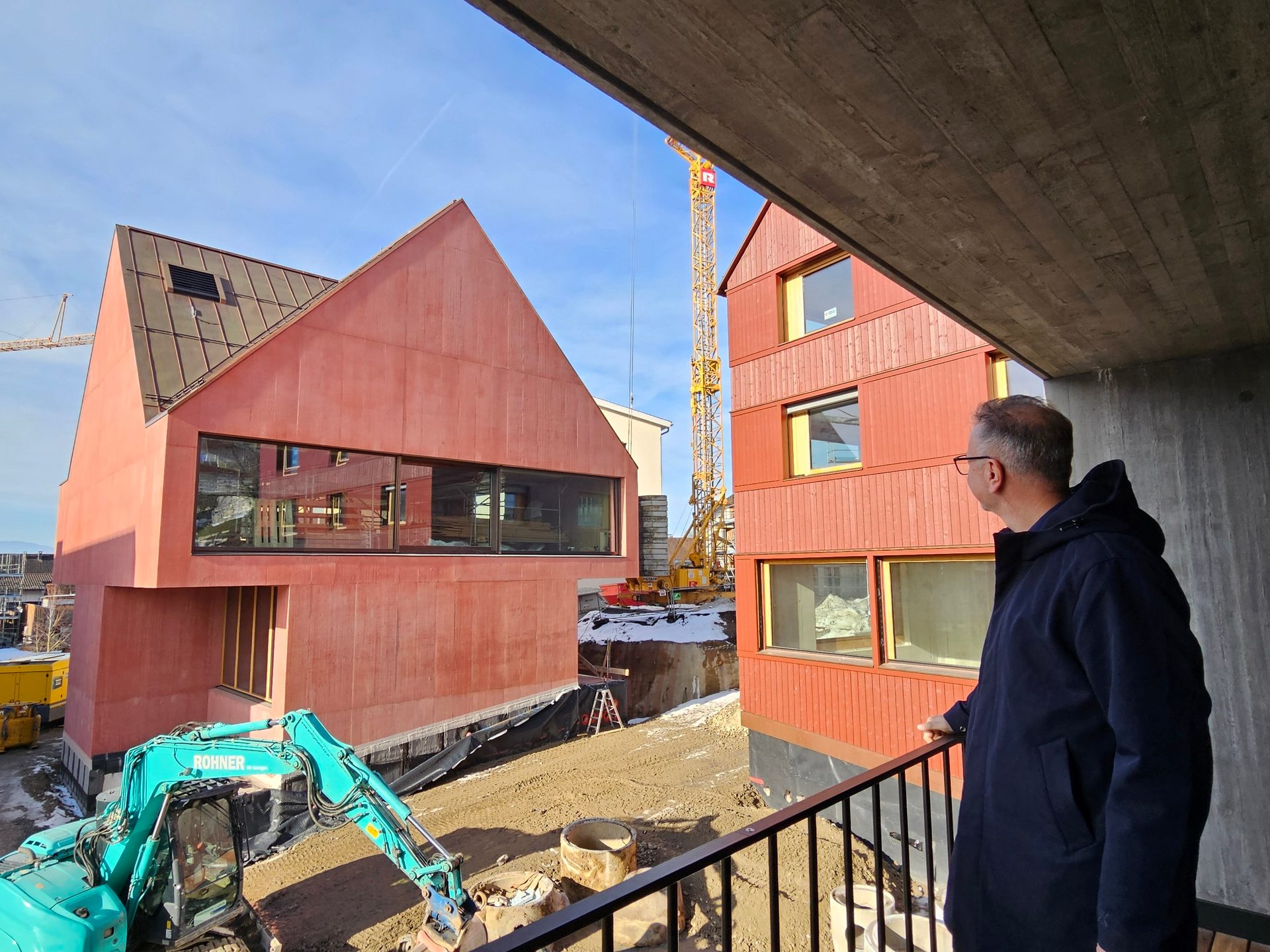 Man on a balcony looking at a construction site with red buildings, an excavator, and a crane.