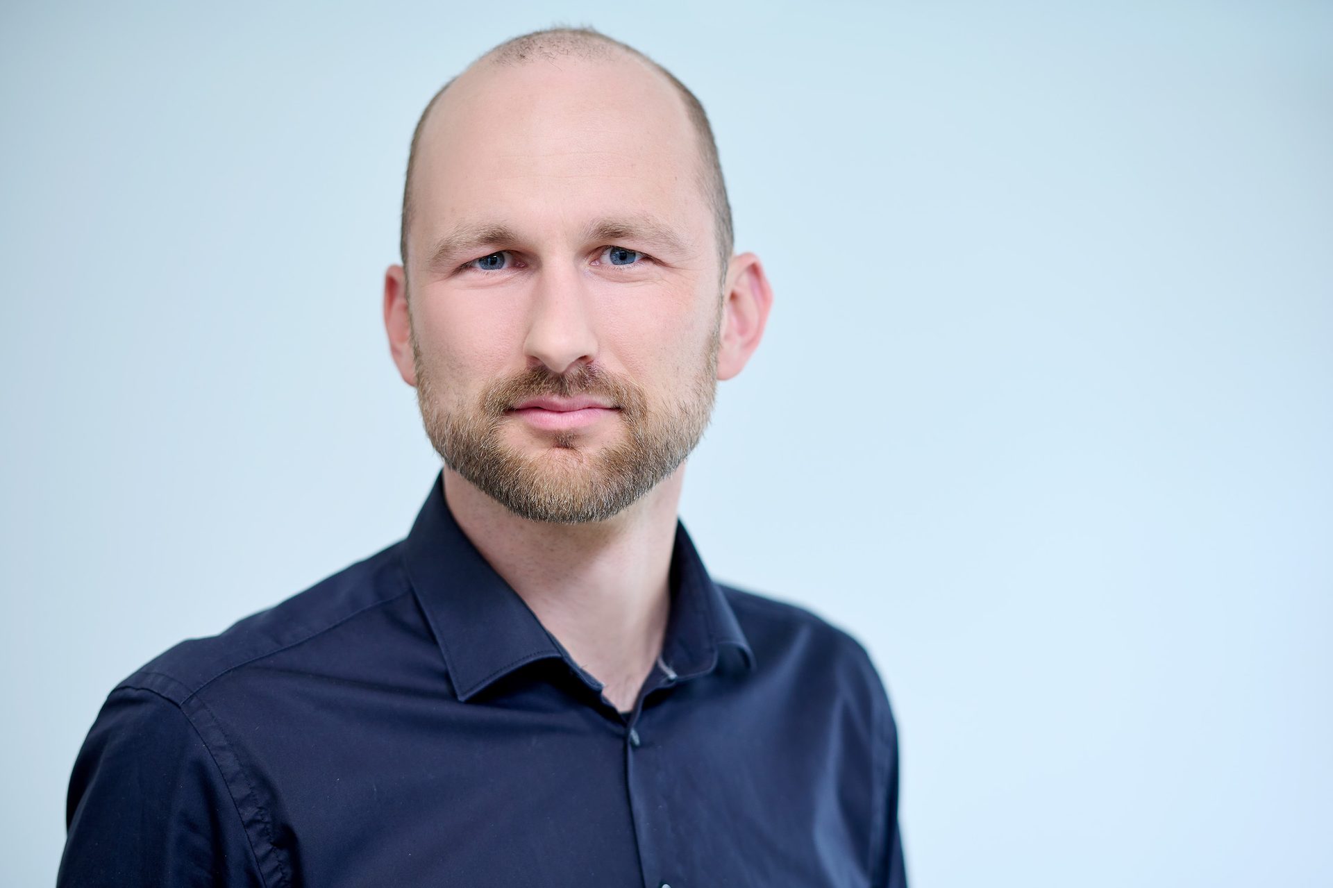 Bald man with blue eyes and beard, dark shirt, looks at viewer.