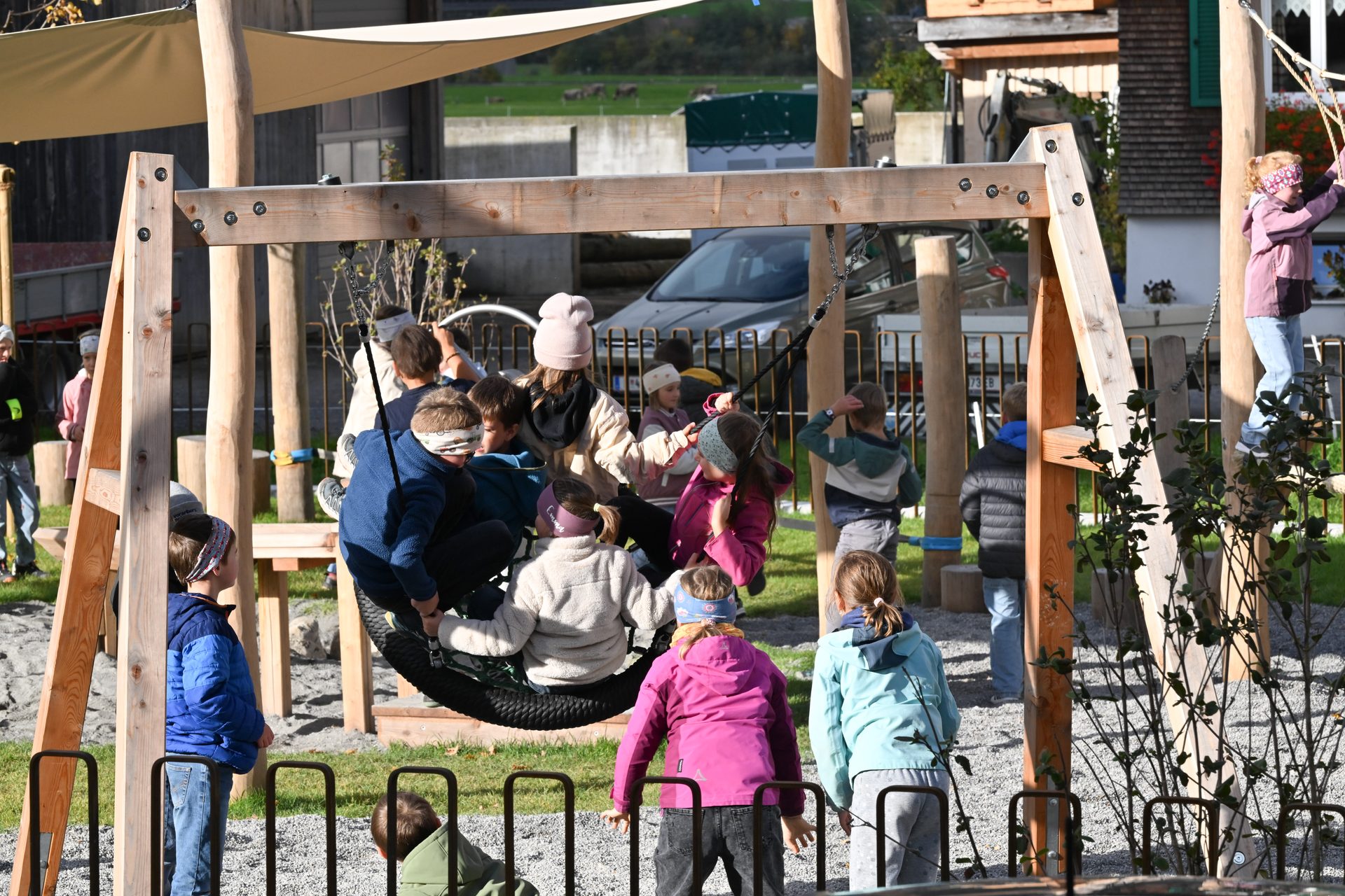 Blindfolded children play on a wooden playground with a large tire swing and climbing ropes.