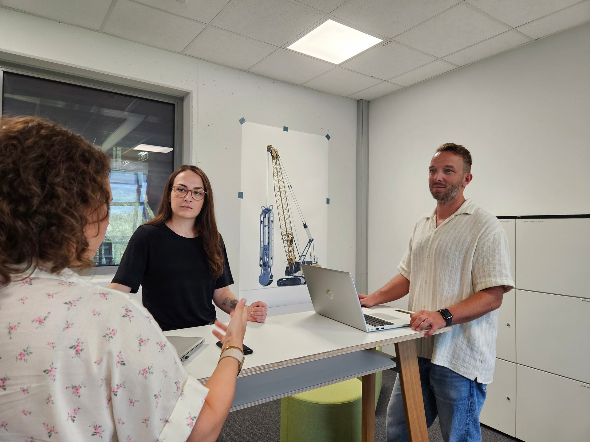 Three people discuss around a laptop at a standing table; a crane poster on the wall.
