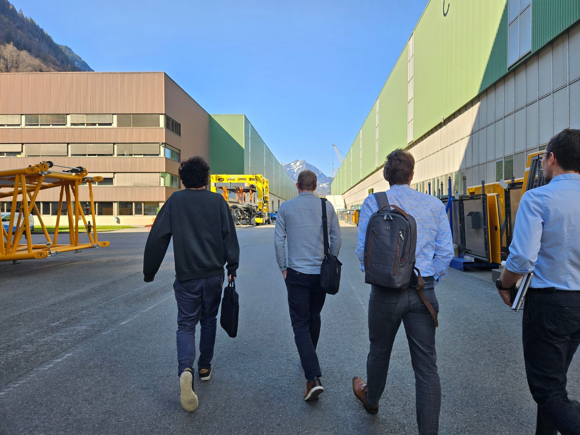 Four men walk along an asphalt path between large industrial buildings toward construction equipment.