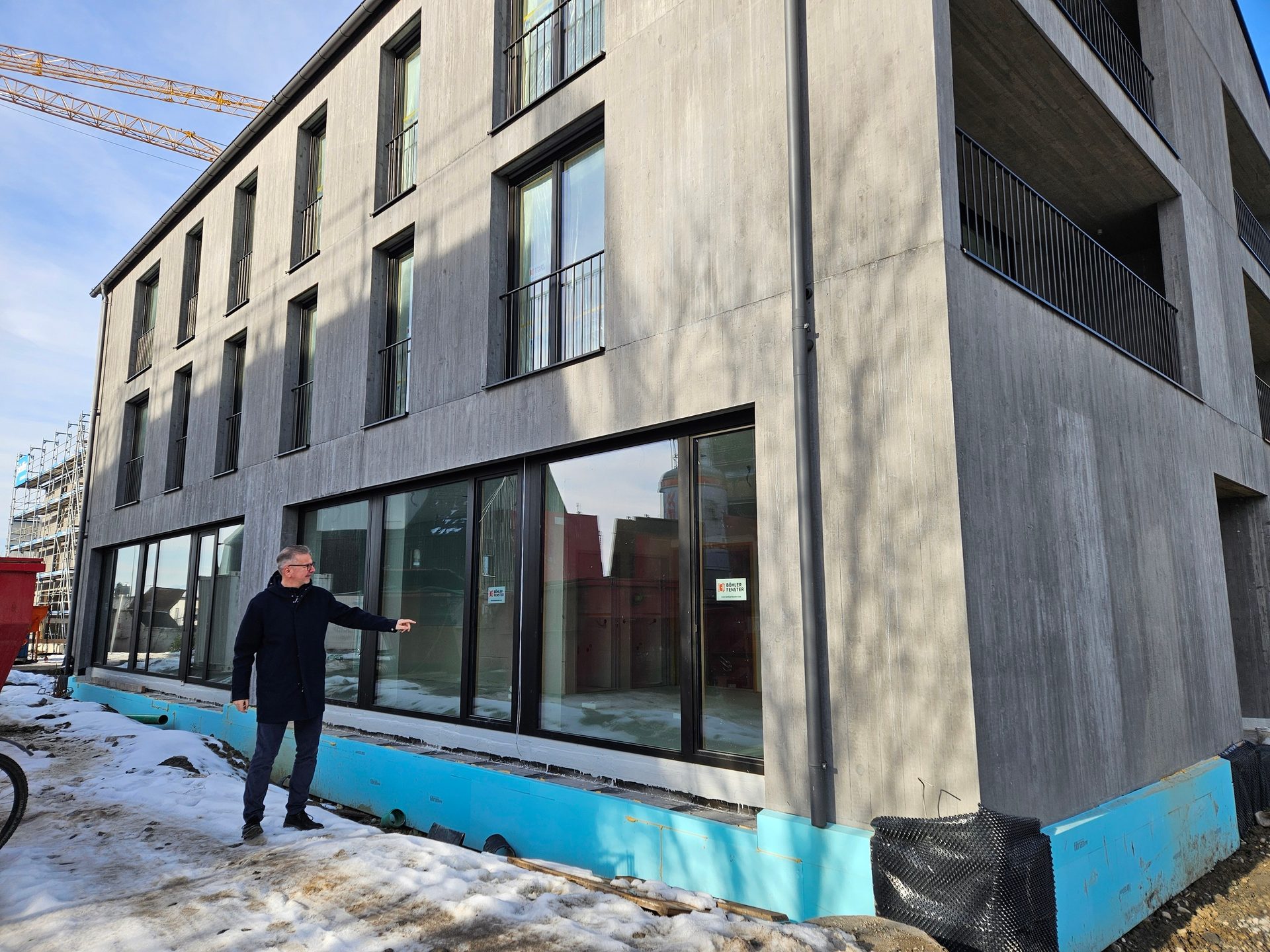 Man pointing at a modern concrete building under construction, with snow and a crane visible.