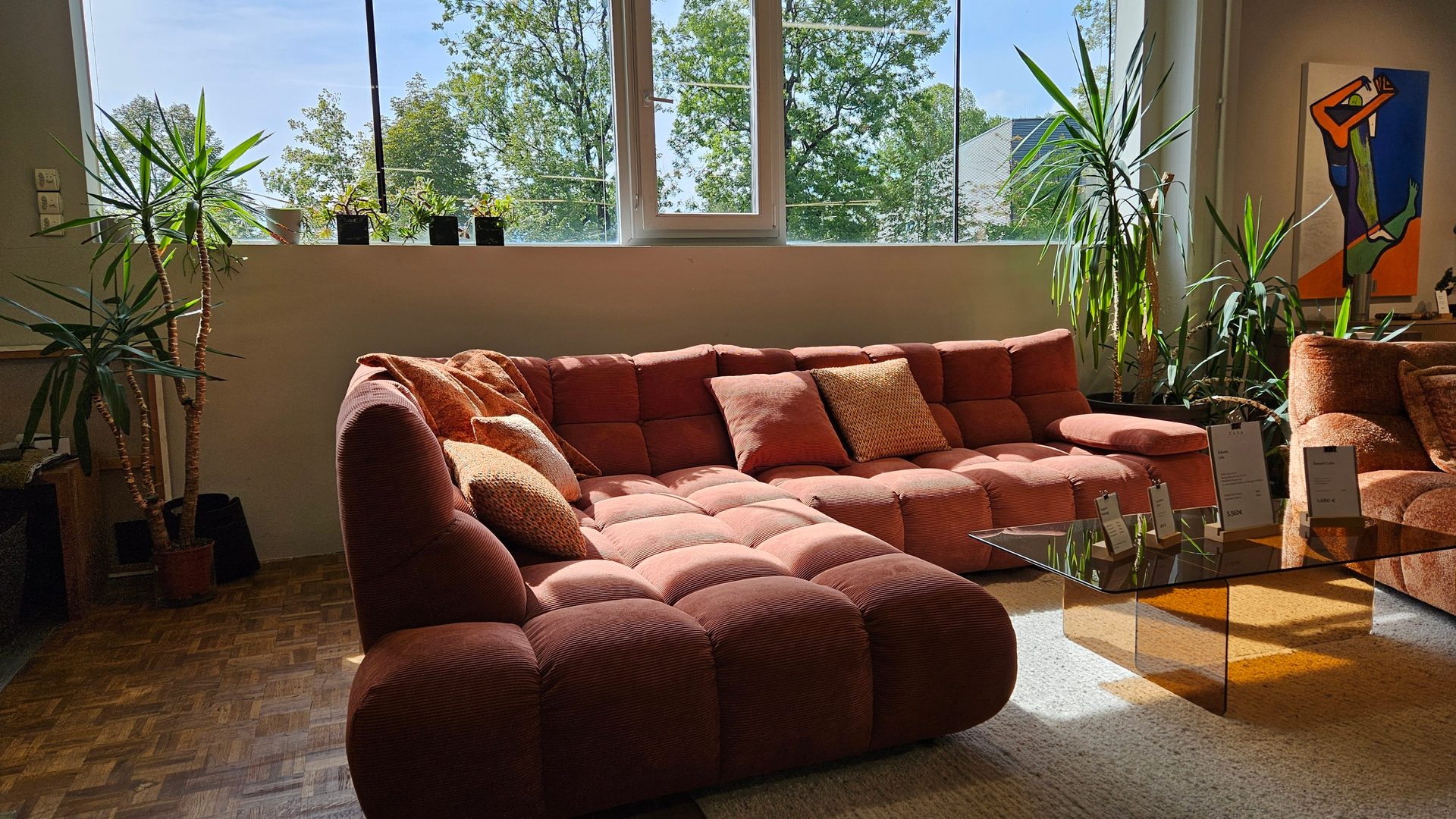 A spacious living room featuring a large reddish-brown sectional sofa, tall plants, and a window with green trees.