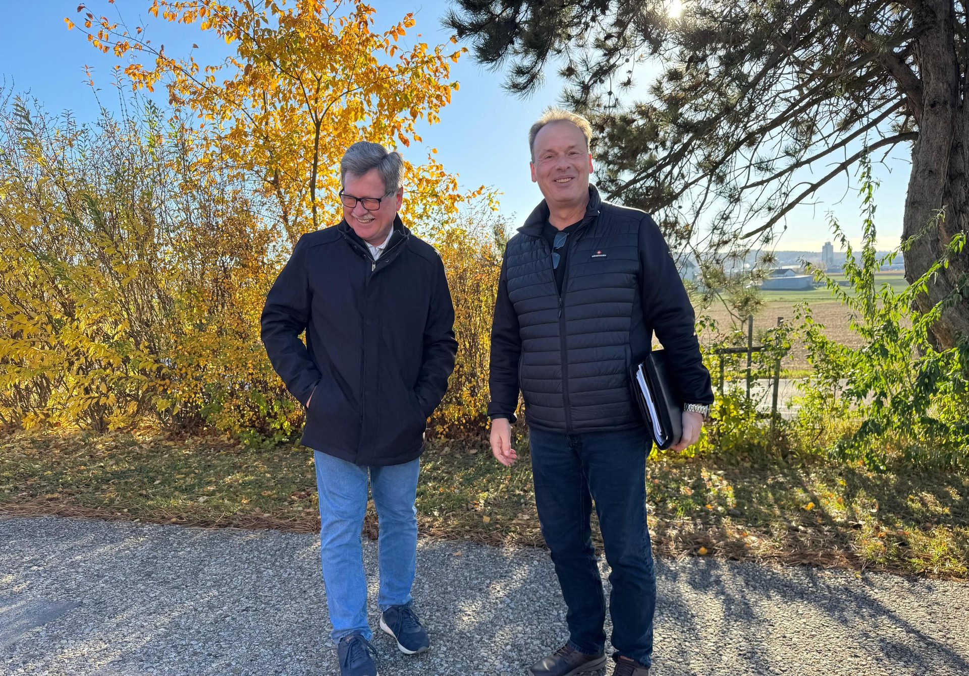 Two smiling men outdoors with vibrant autumn trees.