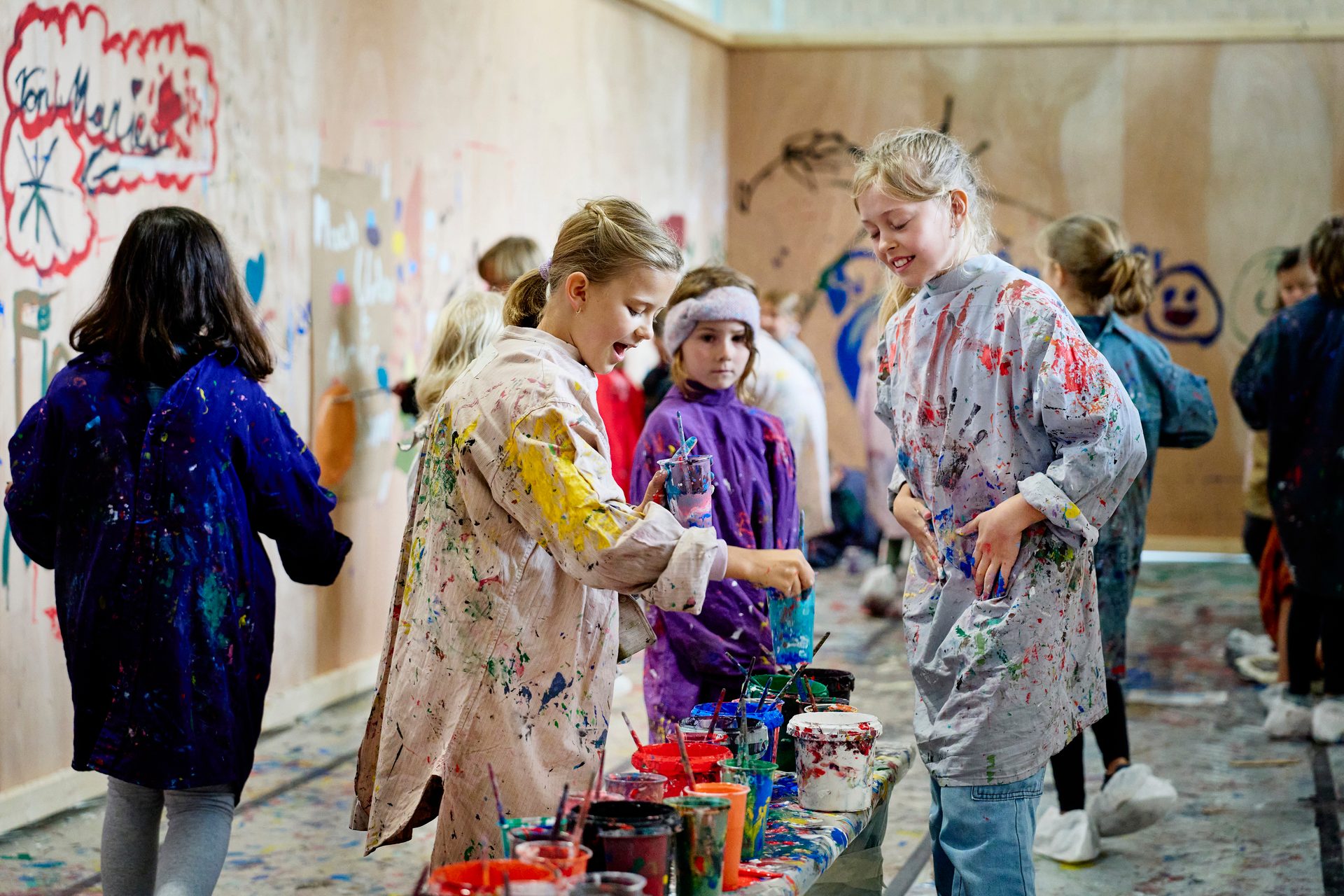 Children in paint-splattered smocks enthusiastically create art in a vibrant classroom.