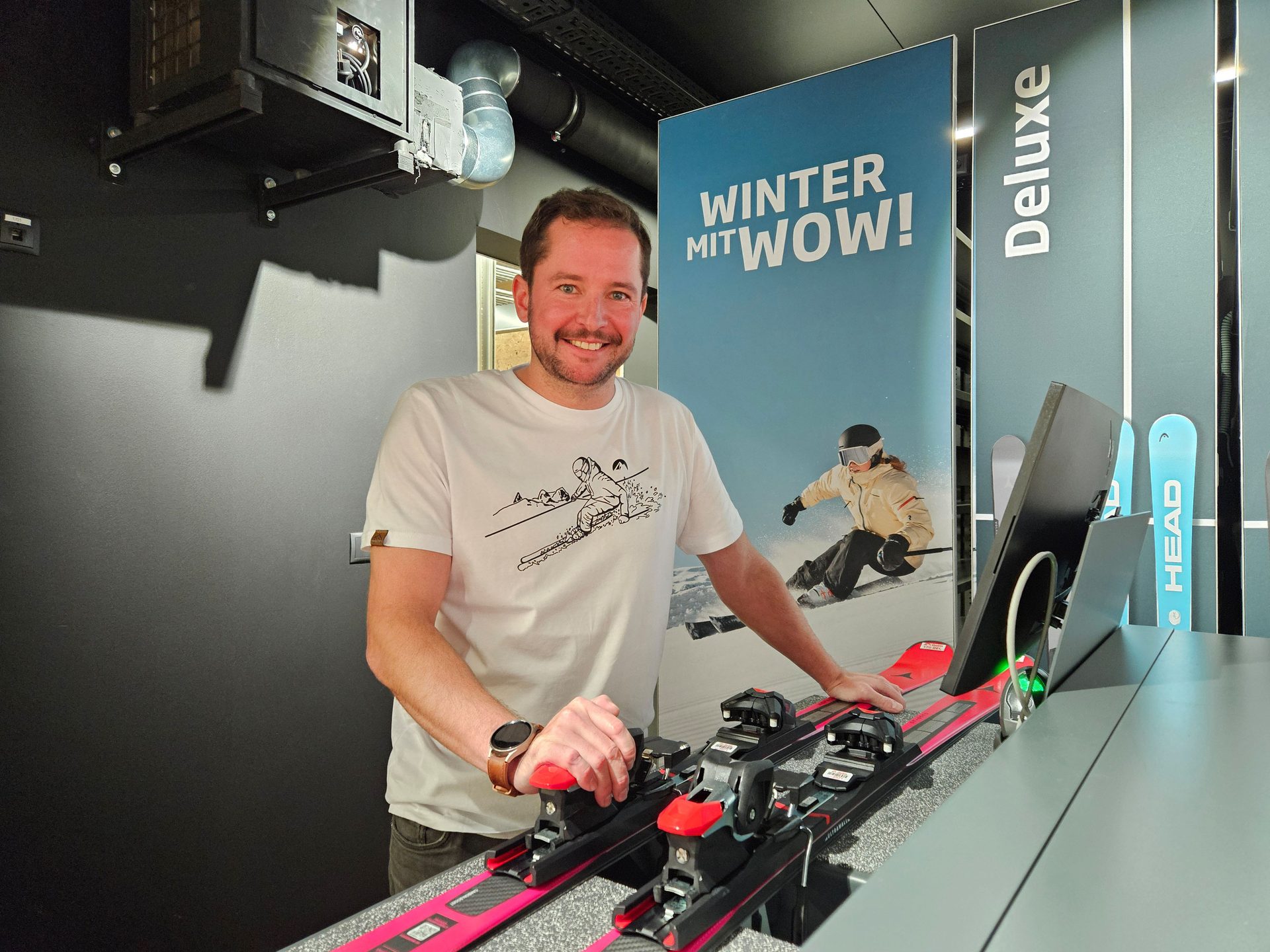 Smiling man holding pink skis in a shop with winter promotional banners.