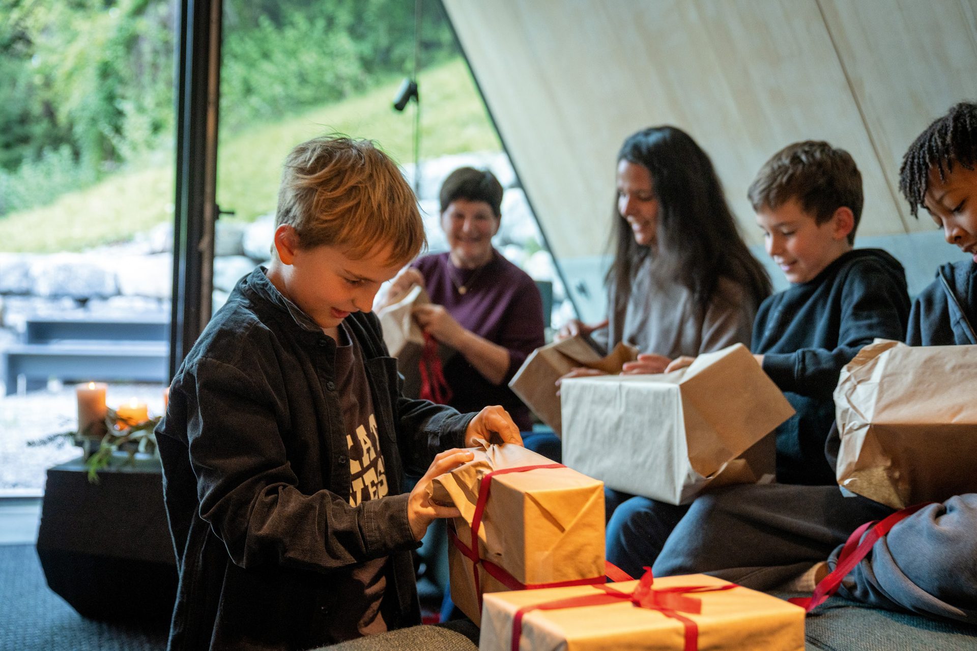 Children and adults happily unwrap brown paper gifts indoors.