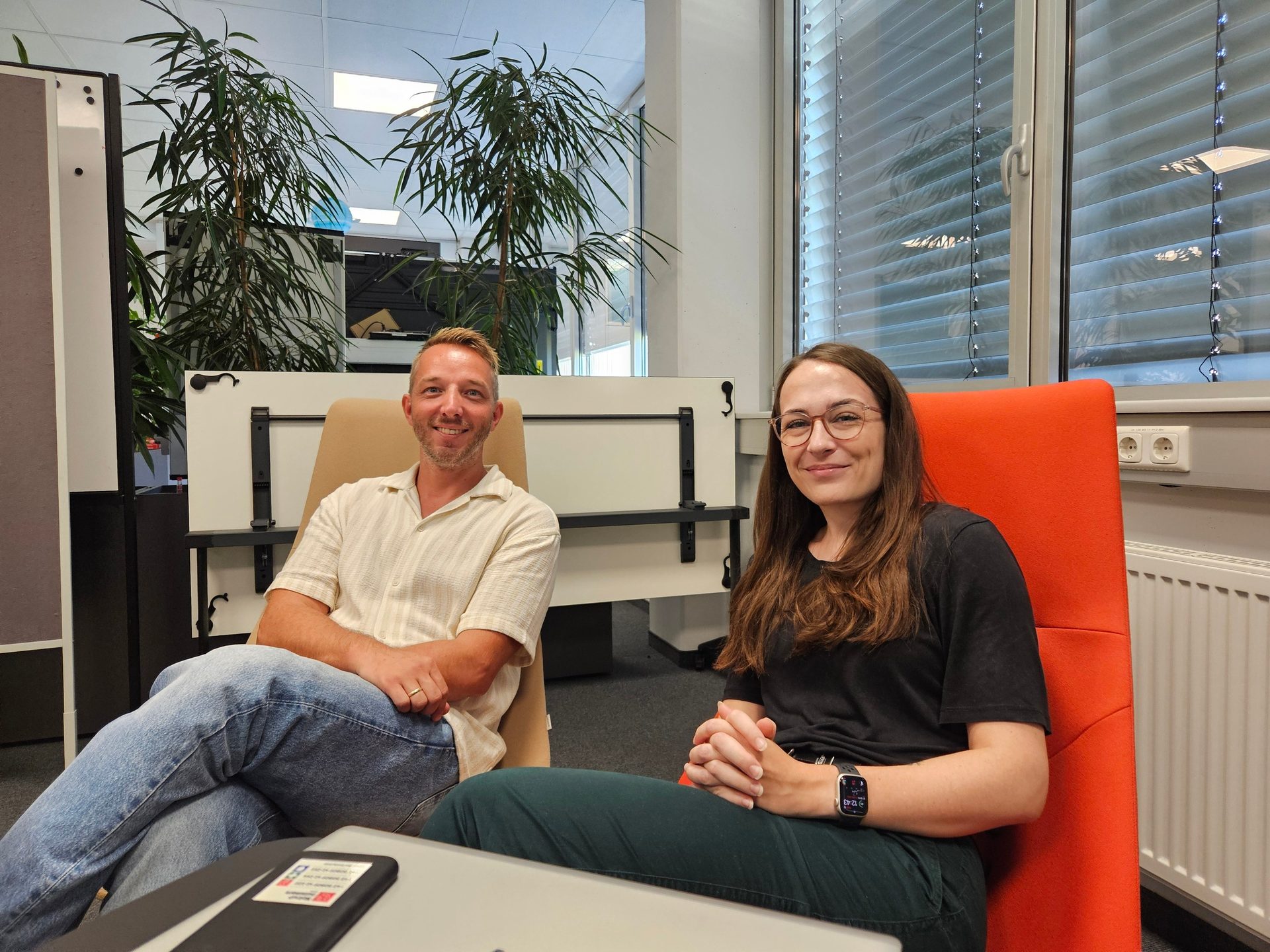 Man and woman sit smiling in an office with plants.