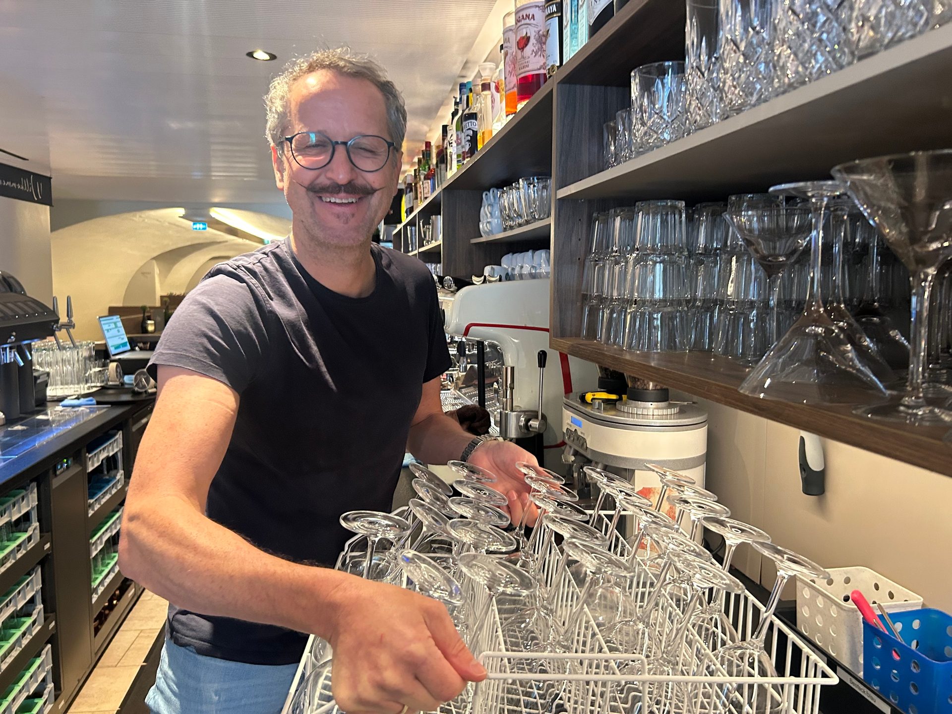 A smiling man with glasses and a mustache holds a rack of clean glasses behind a bar.