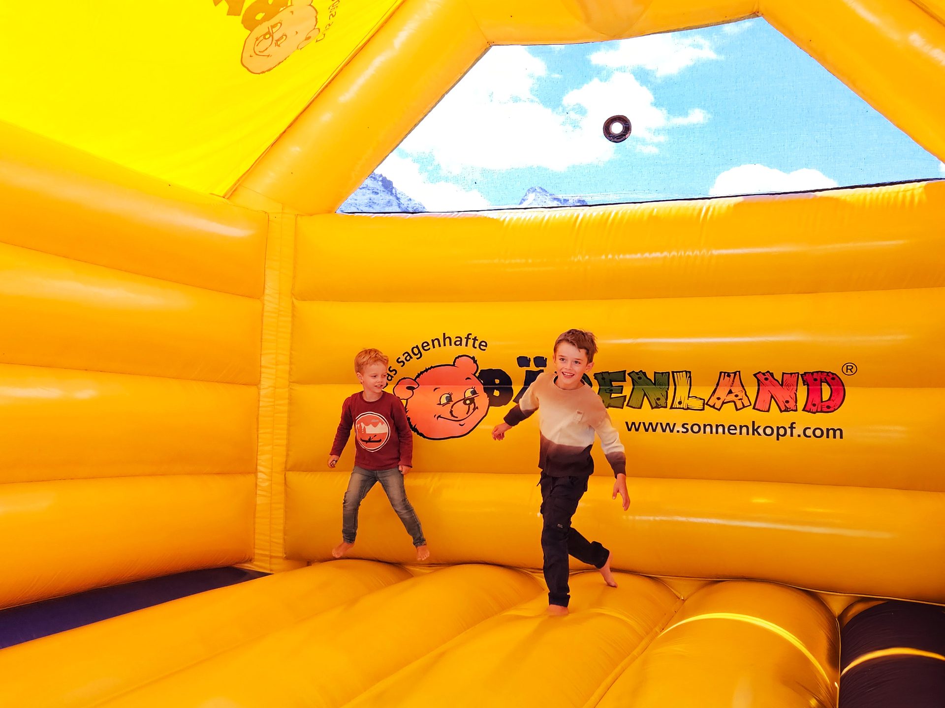 Two boys jump in a yellow bouncy castle with mountains and sky visible.