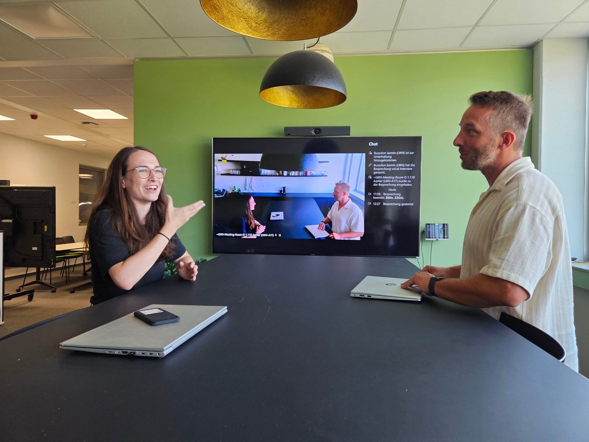 Two people at a table in an office room, with a screen showing a video meeting and chat.