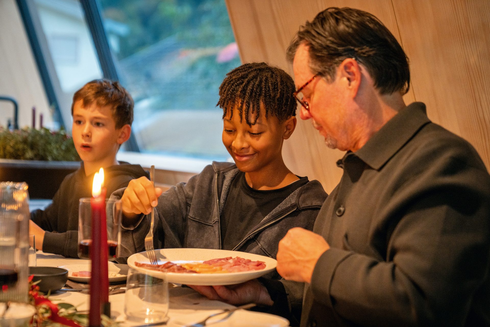 A young person with dreadlocks holds a food plate with a fork, an older man watches intently at a table.