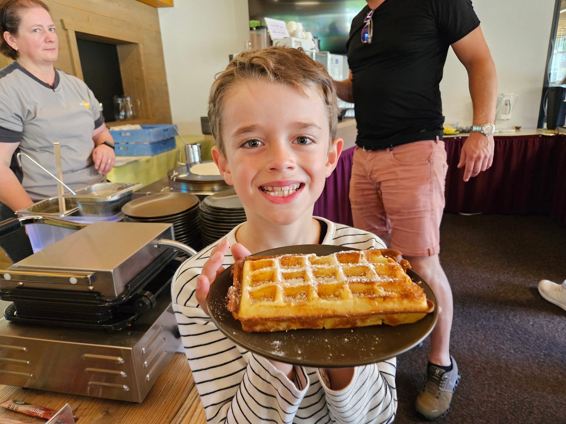 A smiling boy proudly holds a fresh waffle topped with powdered sugar at a breakfast station.