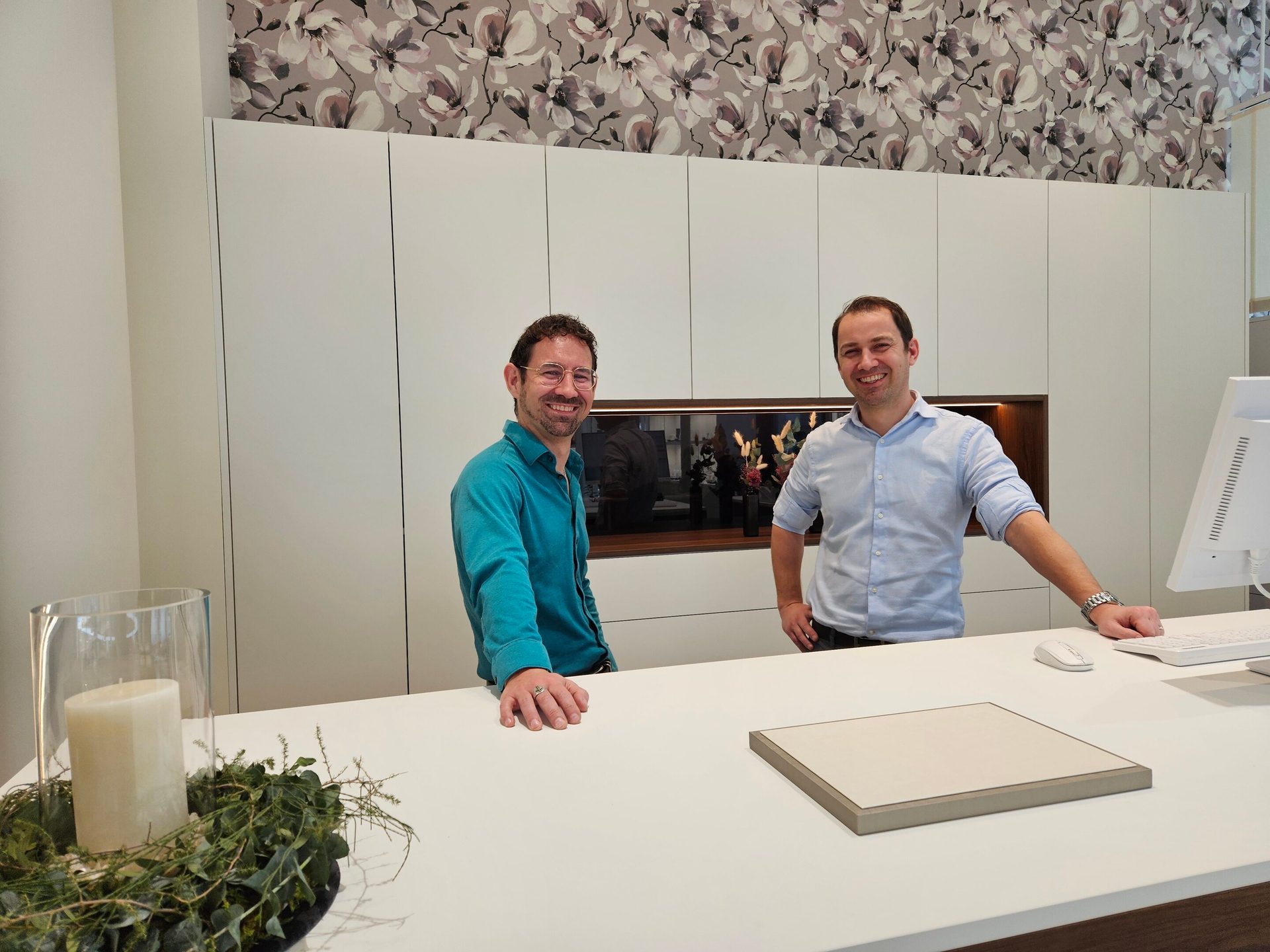 Two smiling men behind a white counter with floral wallpaper.