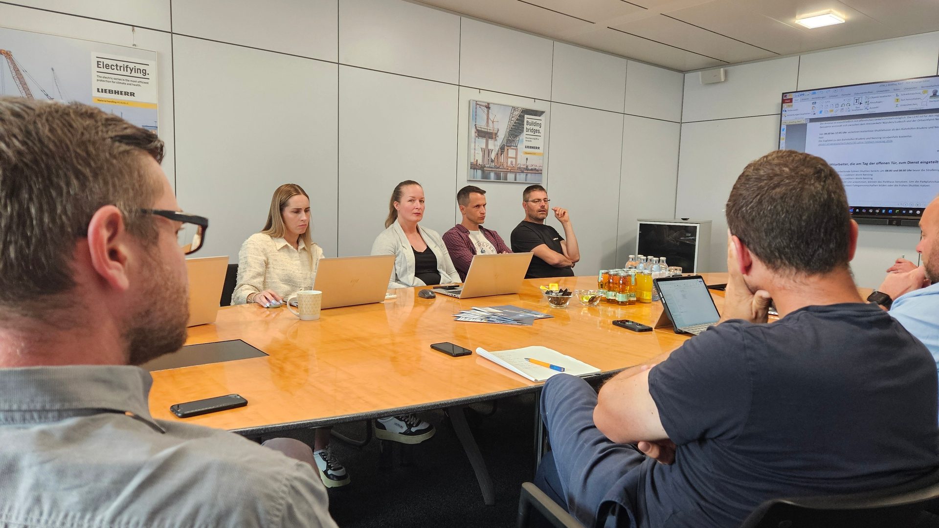 People in a meeting room, sitting around a wooden table with laptops. A large screen is in background.