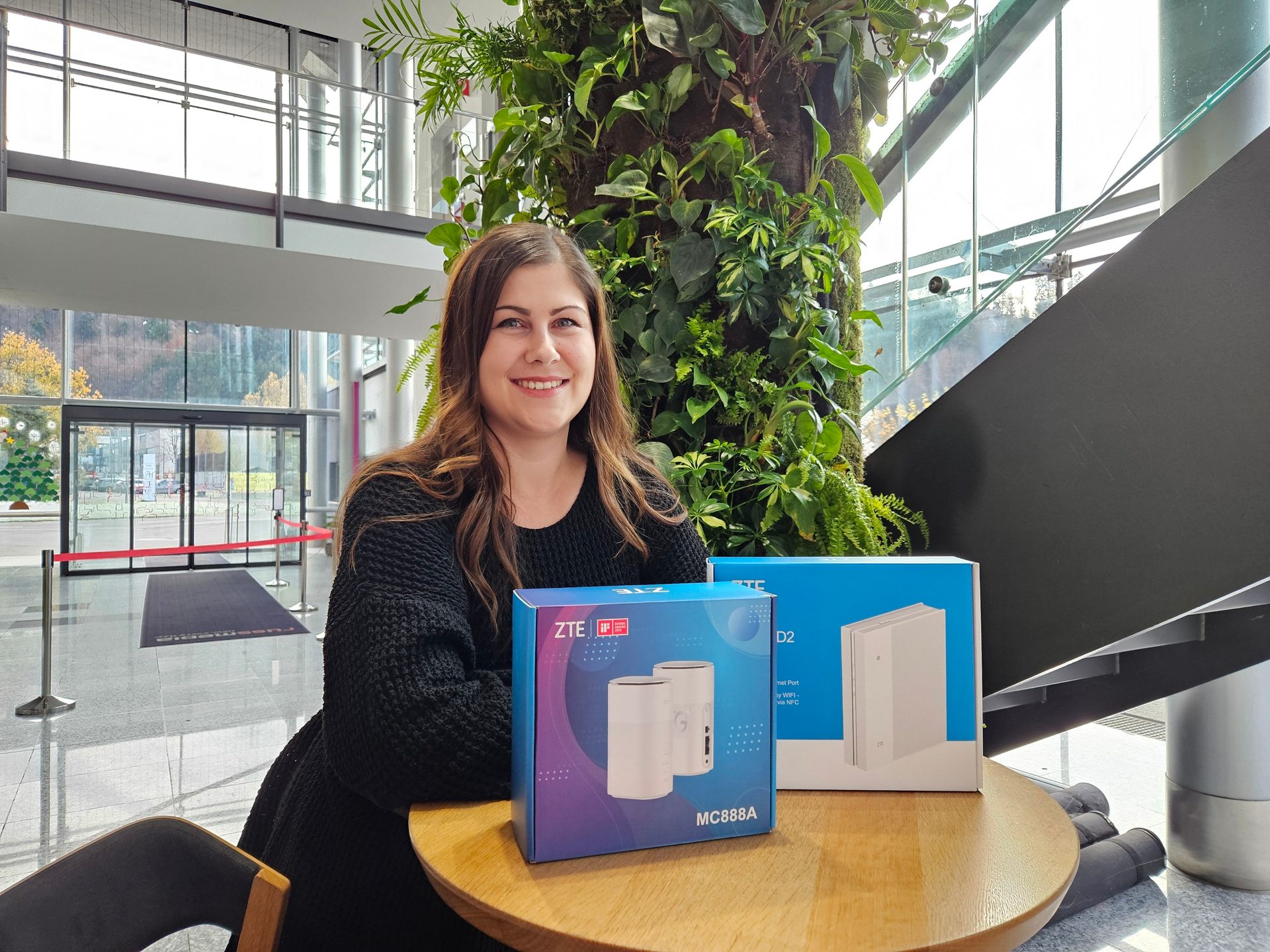 Smiling woman with two ZTE router boxes on a table, in a modern building with a vertical garden.