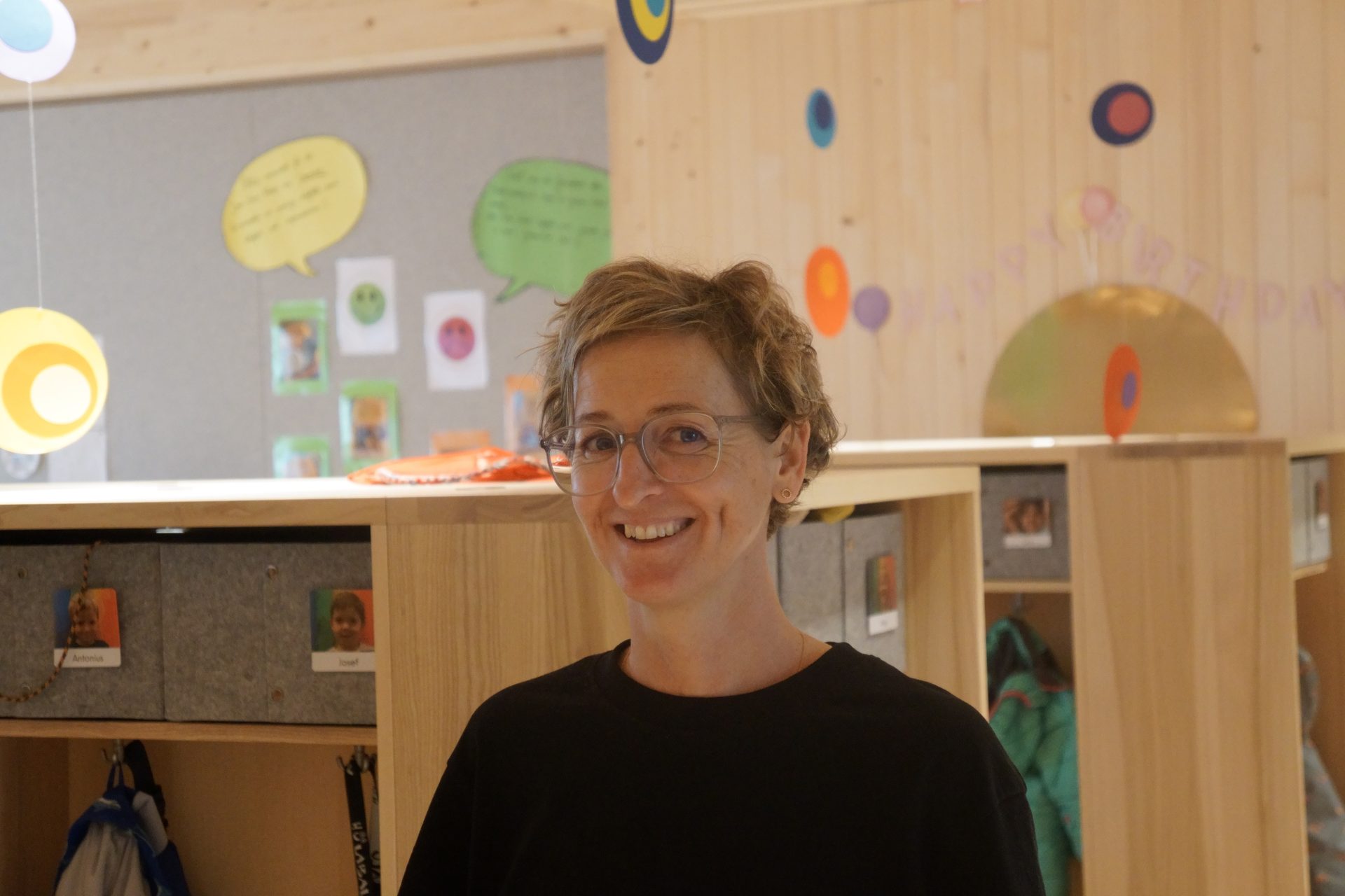 Smiling woman in glasses in a cheerful classroom with colorful decorations and cubbies.