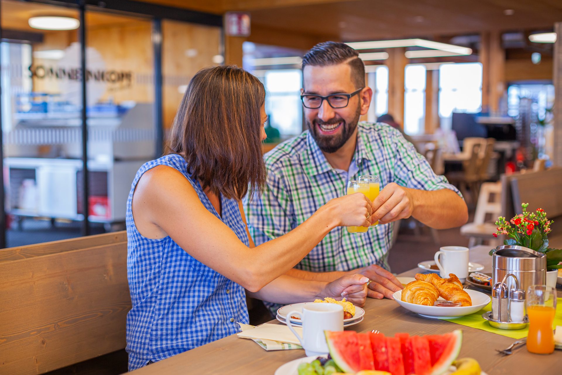 A happy couple smiles while toasting orange juice glasses during a breakfast meal with fresh food.