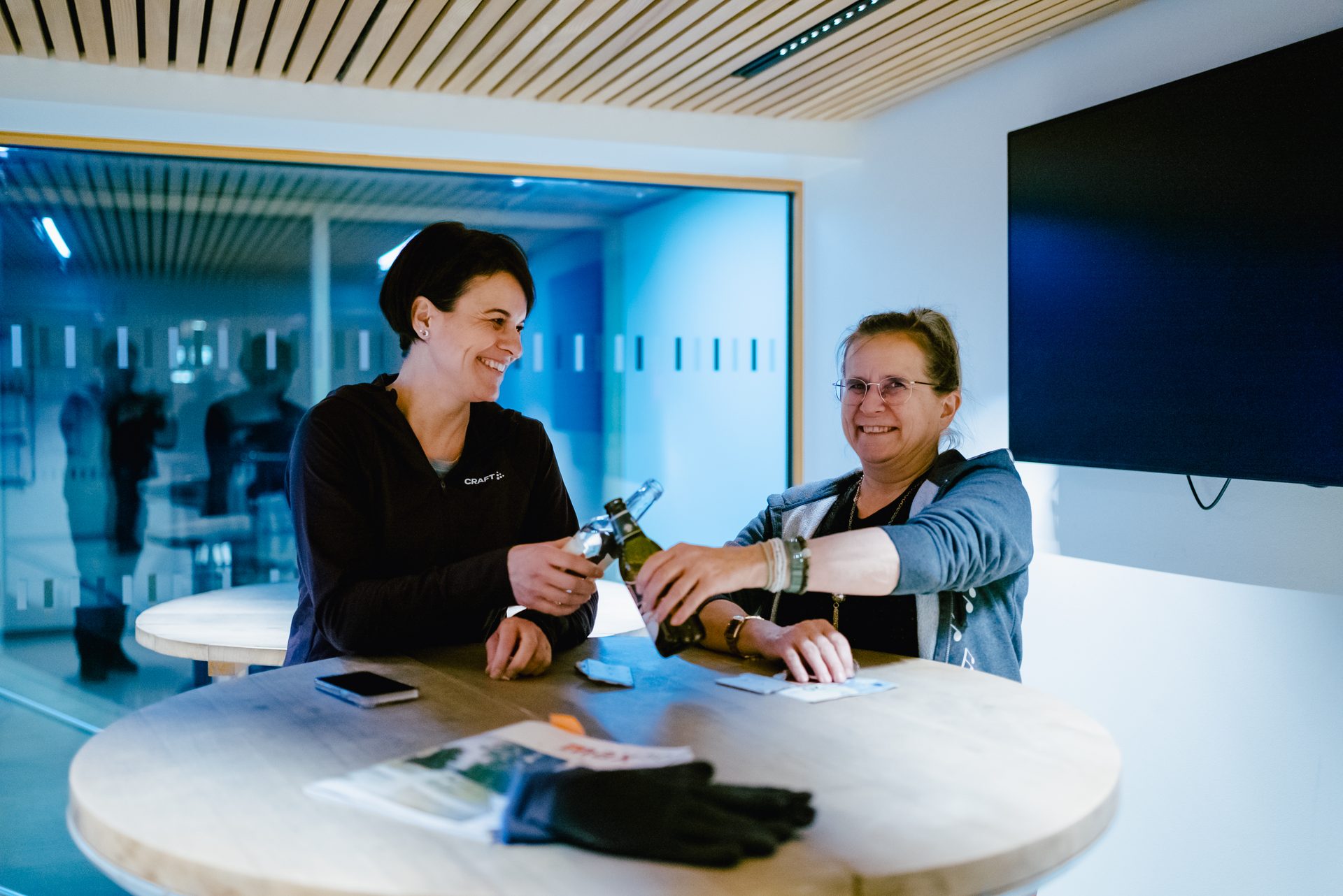 Two women celebrating with drinks at a table in a modern office or common area, smiling happily.