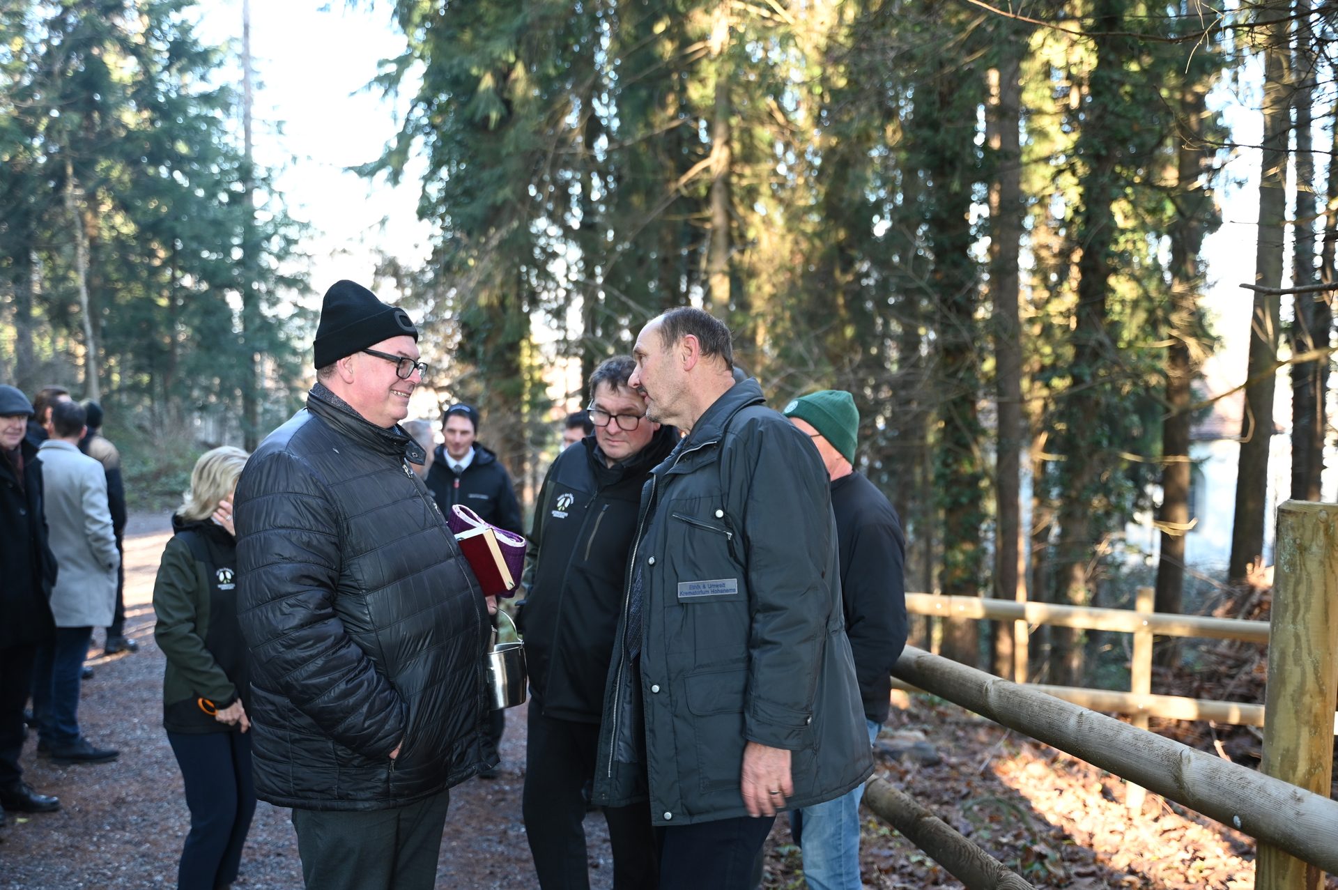 A group of people gathered in a sunny forest, with tall trees and a wooden railing visible.