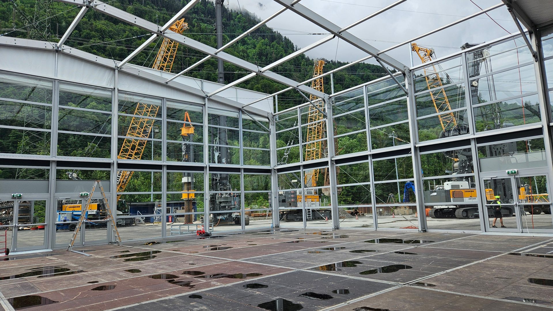 Interior of a transparent construction tent, showing heavy machinery, mountains, and wet floor.