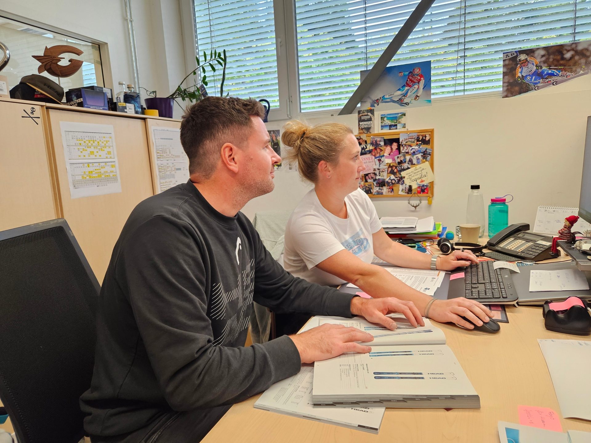 Two colleagues work at an office desk; one uses a computer, the other reviews a HEAD catalog.