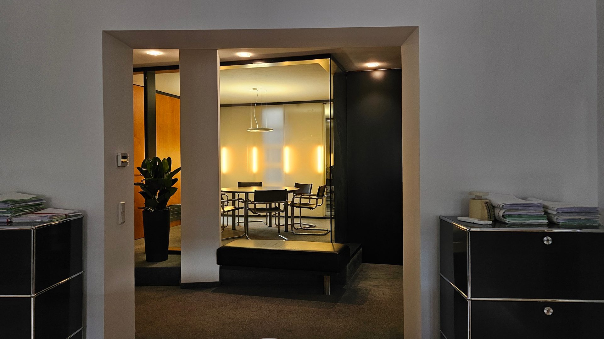 Modern conference room with round table, chairs, and vertical lights, viewed from an office hallway.