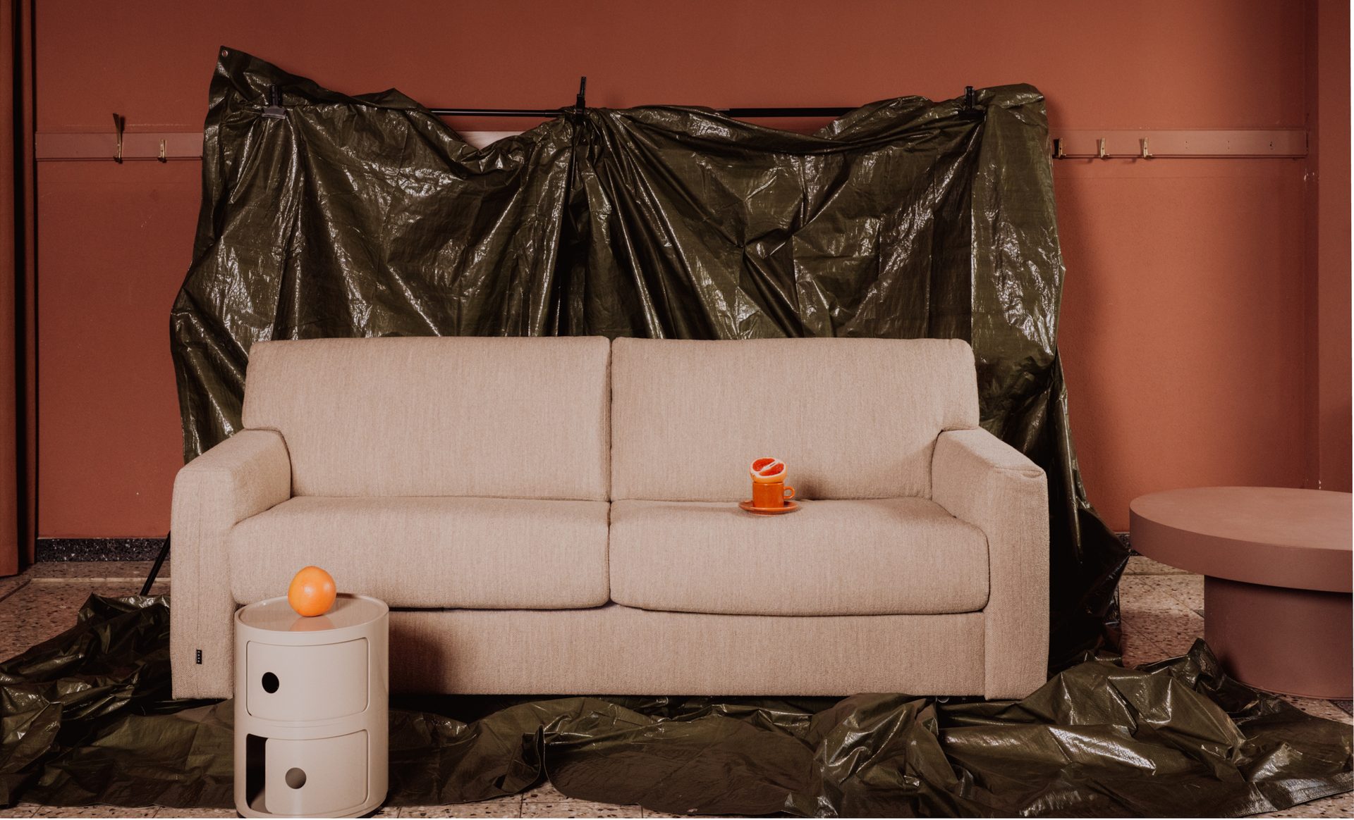 Beige sofa, two side tables, and orange items in a room with a red wall and draped tarp.