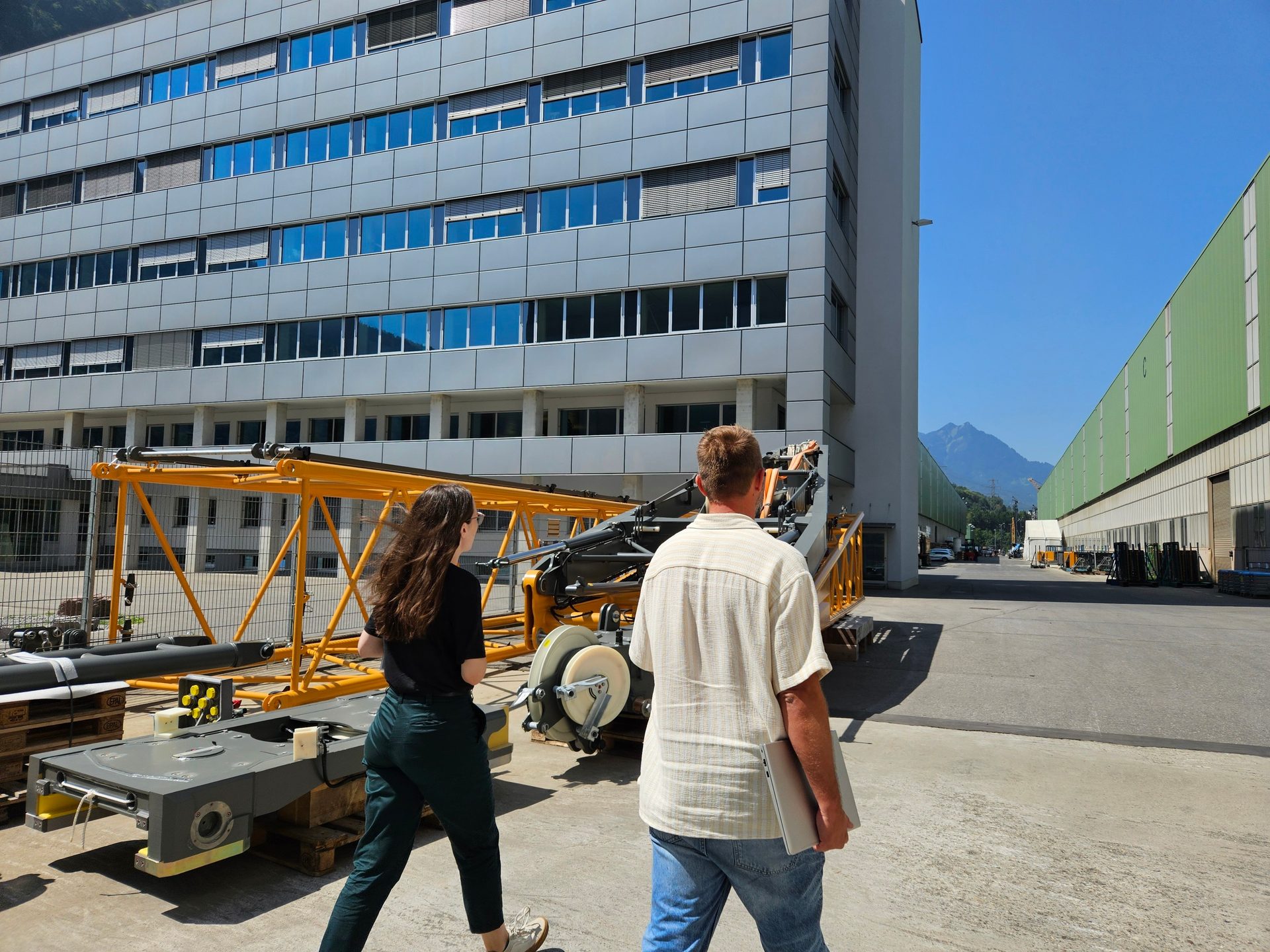 Two people walk past large industrial machinery and buildings under a clear sky.