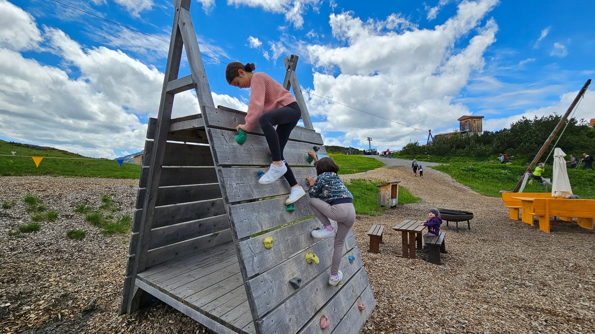 Two girls climb a wooden structure on an outdoor mountain playground under a blue sky.