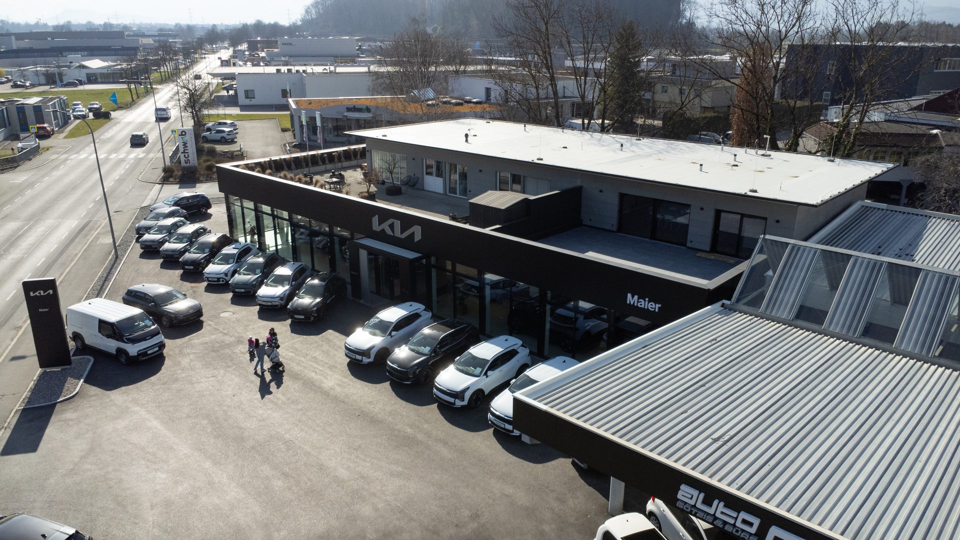 Aerial view of a Kia car dealership with new vehicles parked outside and a family walking.