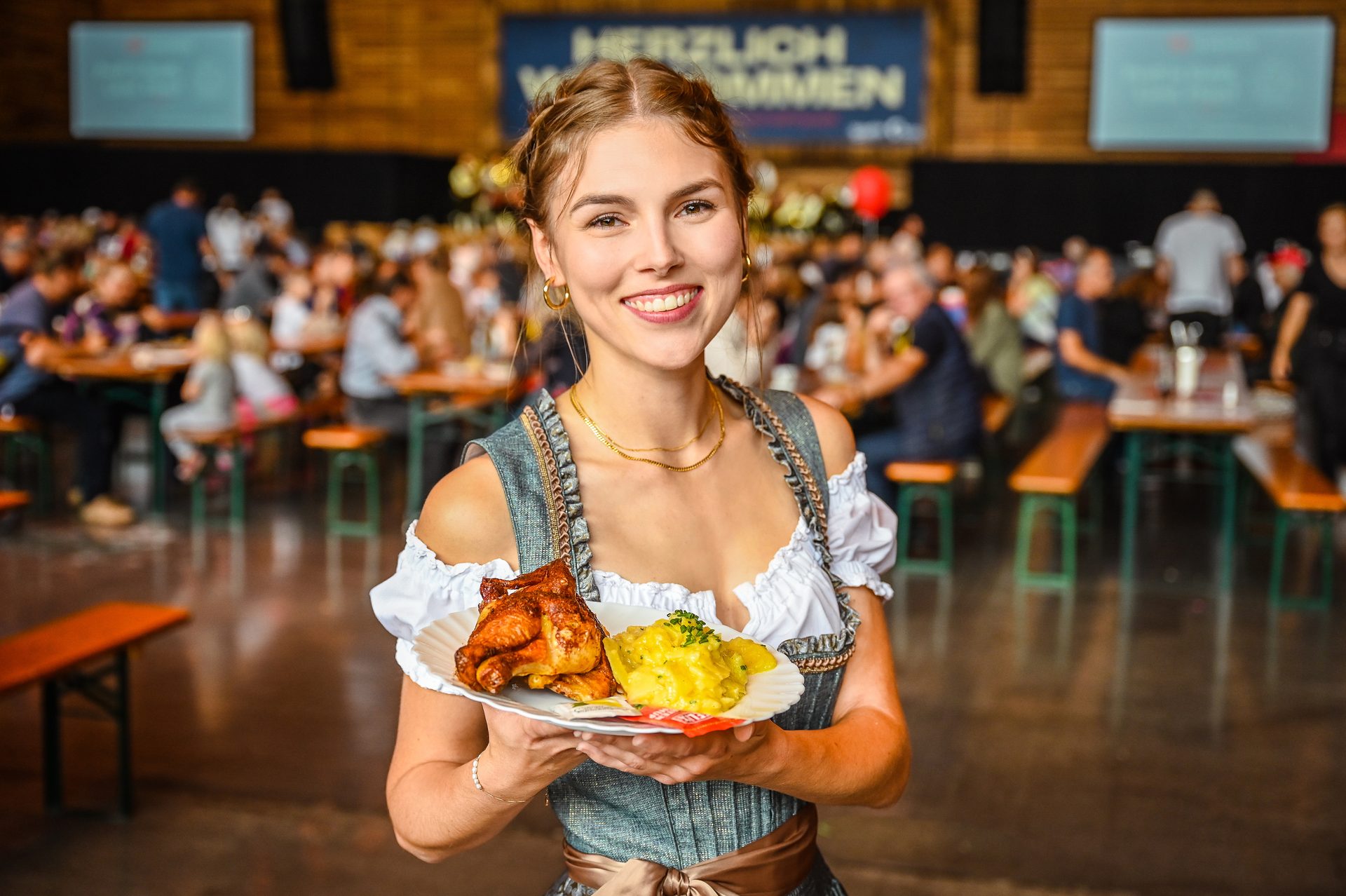 A smiling woman in a dirndl holds a plate with roasted chicken and potato salad in a busy hall.