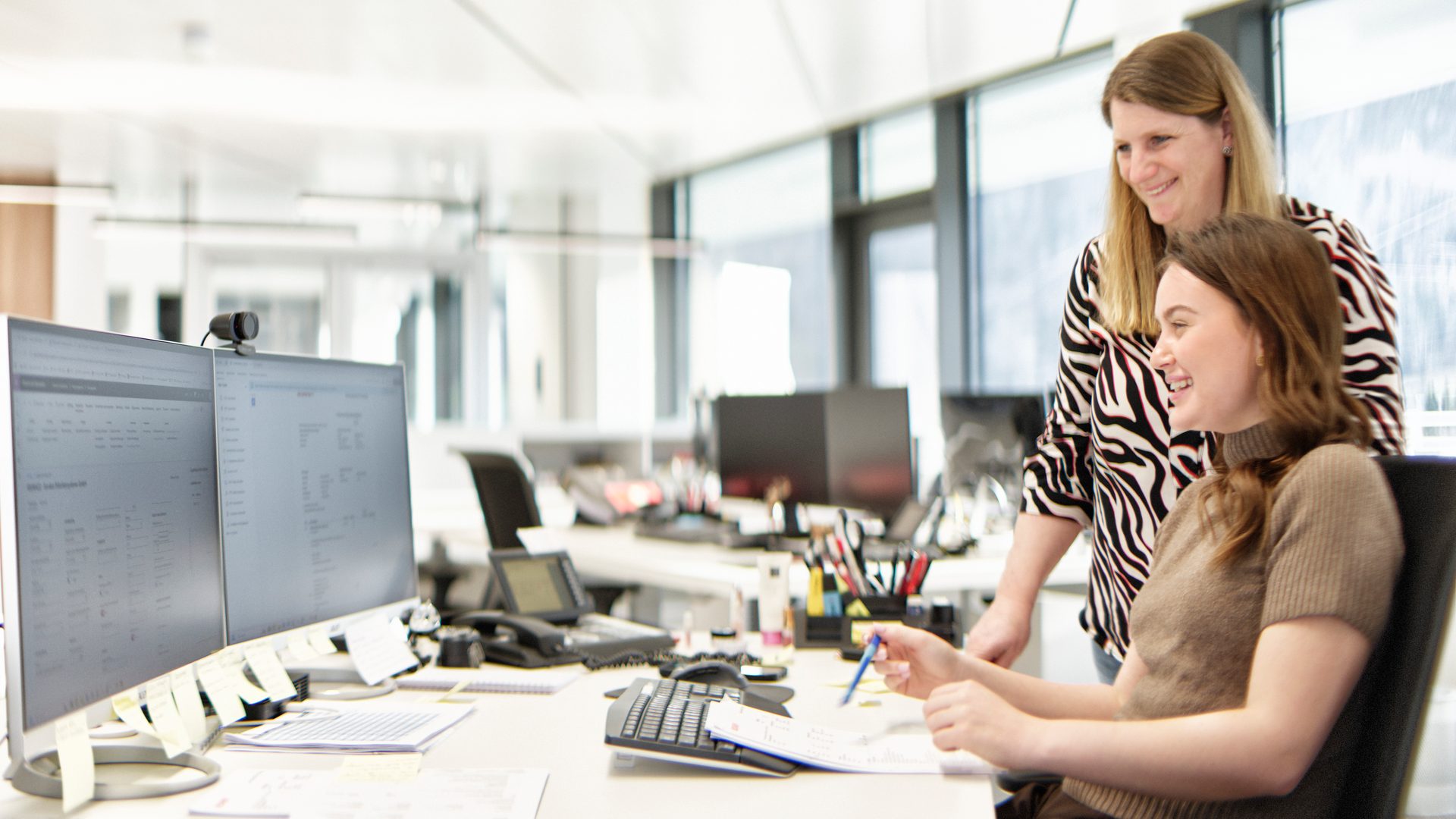 Two women smiling and collaborating at a desk in a bright, modern office environment.